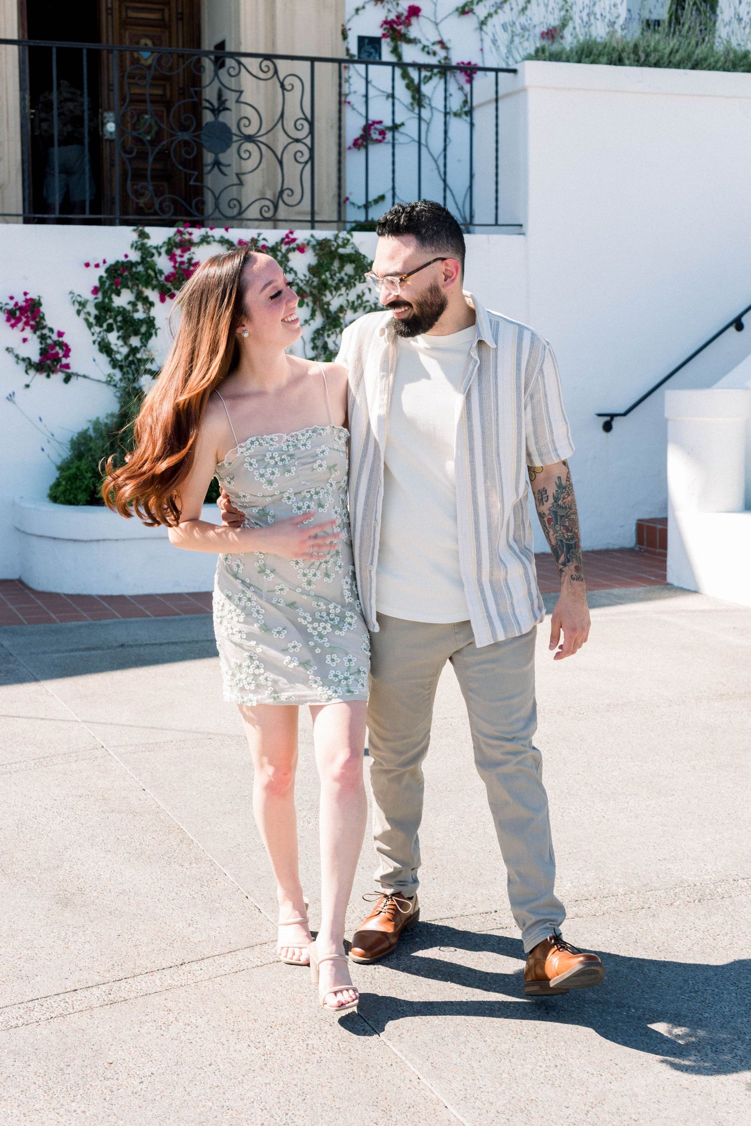 A couple shares a playful moment in casual attire against a white Mediterranean-style building.