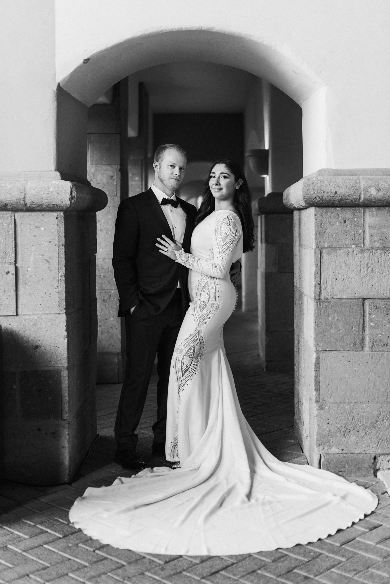 A black and white portrait of newlyweds embracing under a stone archway.