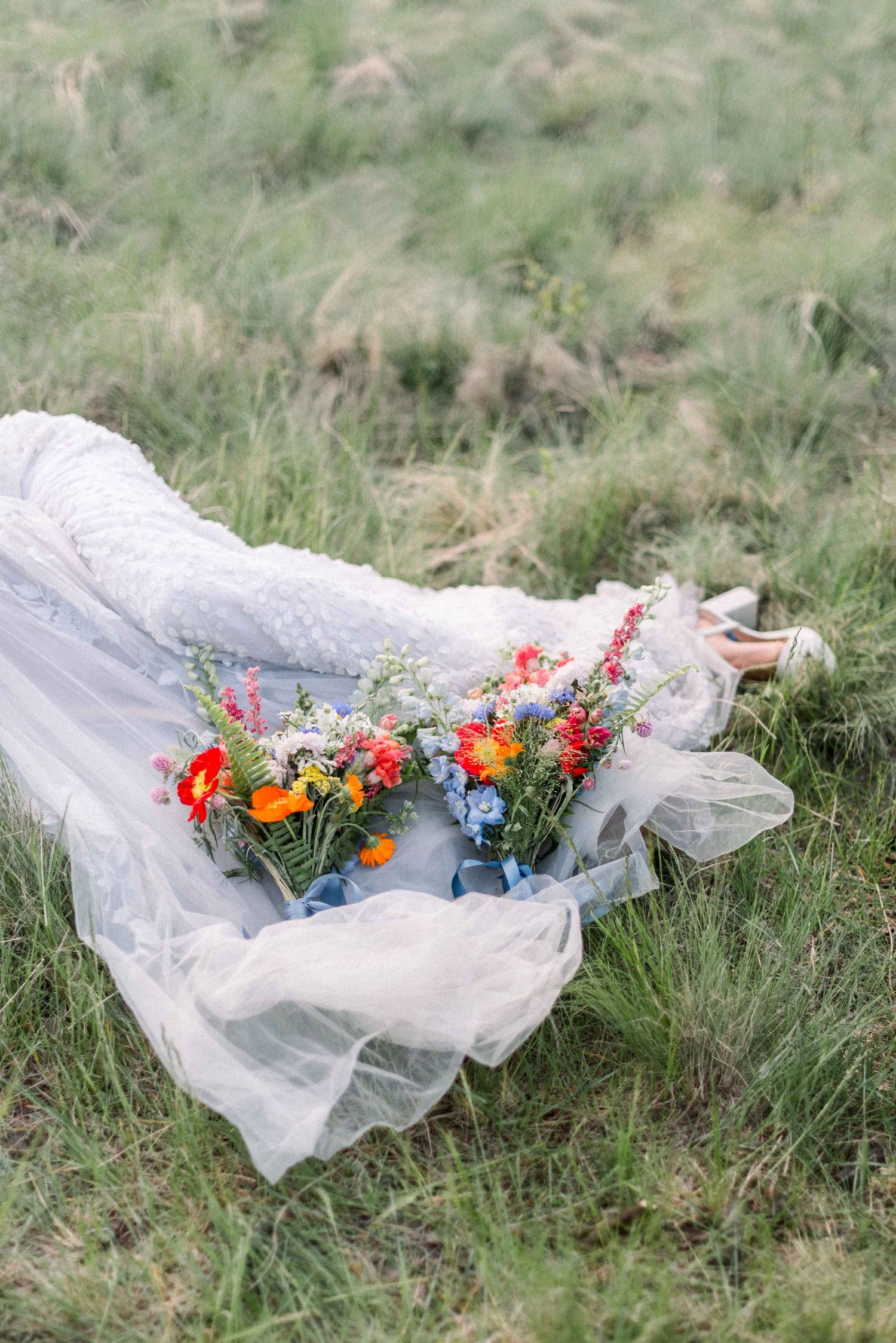 White wedding dress with flowing train and colorful floral bouquet laid out on grassy ground.