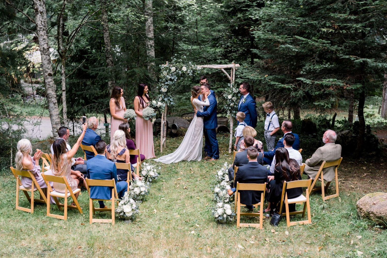 An intimate forest wedding ceremony with guests seated on wooden chairs and floral decorations.