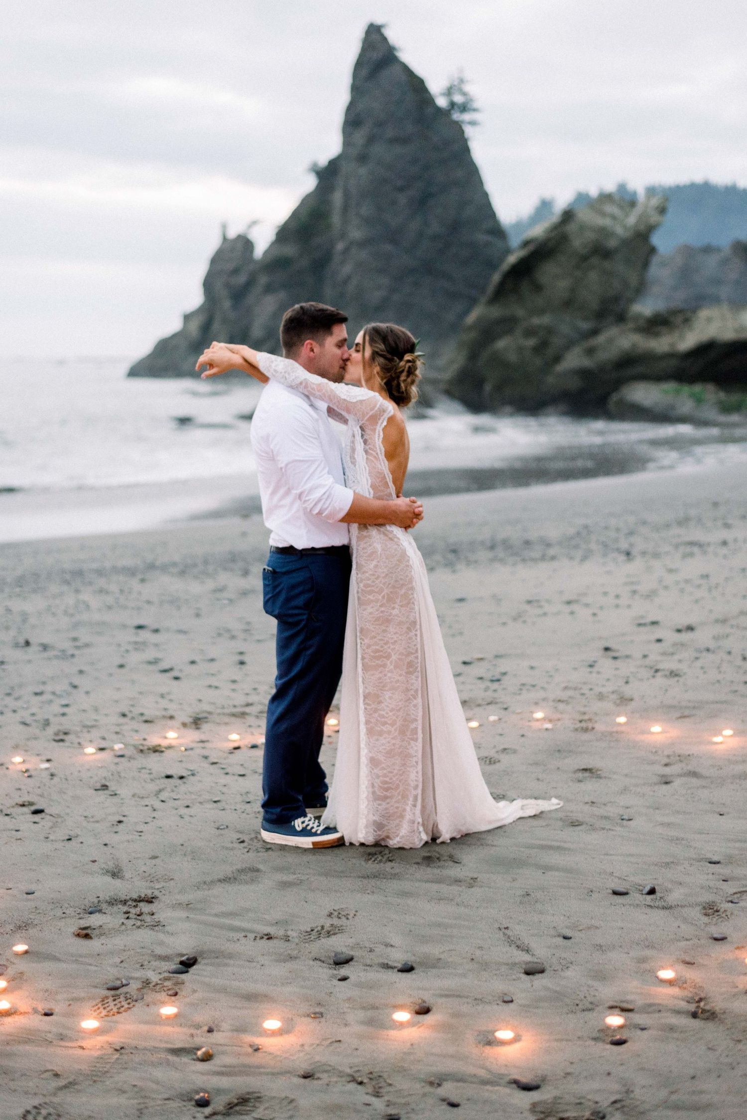 Newlyweds share an intimate dance surrounded by candles on a misty beach at dusk.