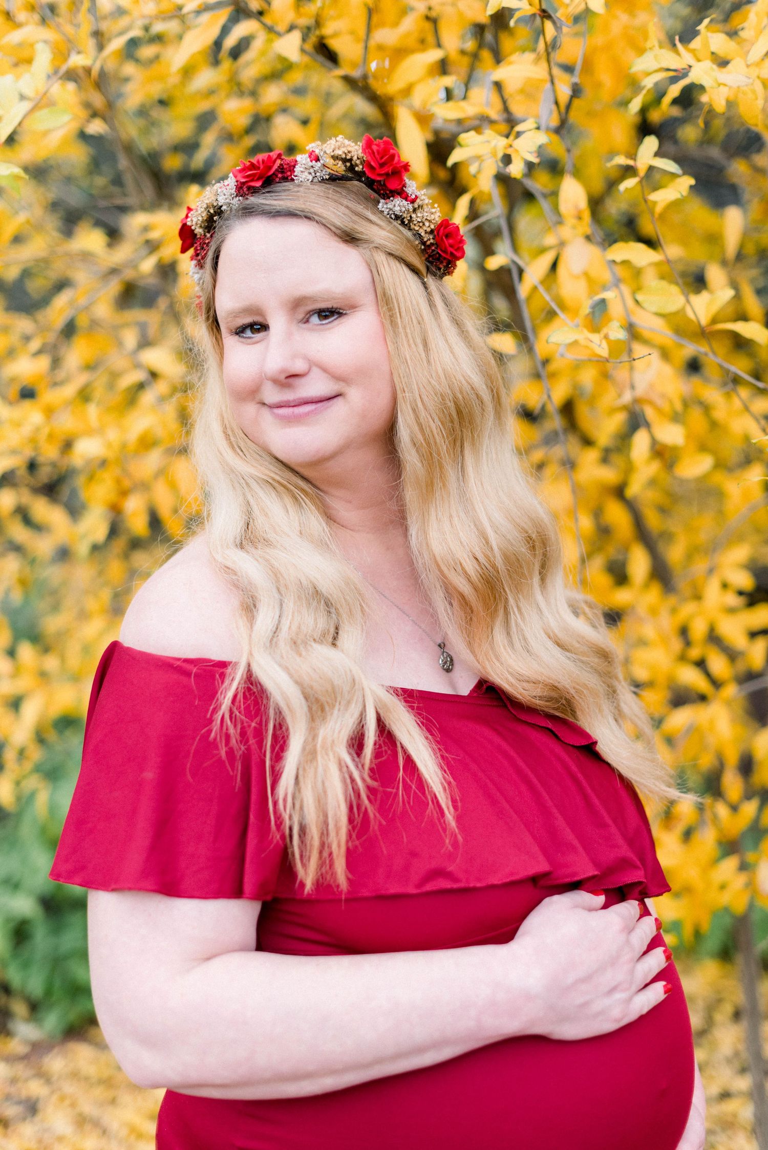 Maternity portrait with floral crown against golden fall leaves wearing red dress.