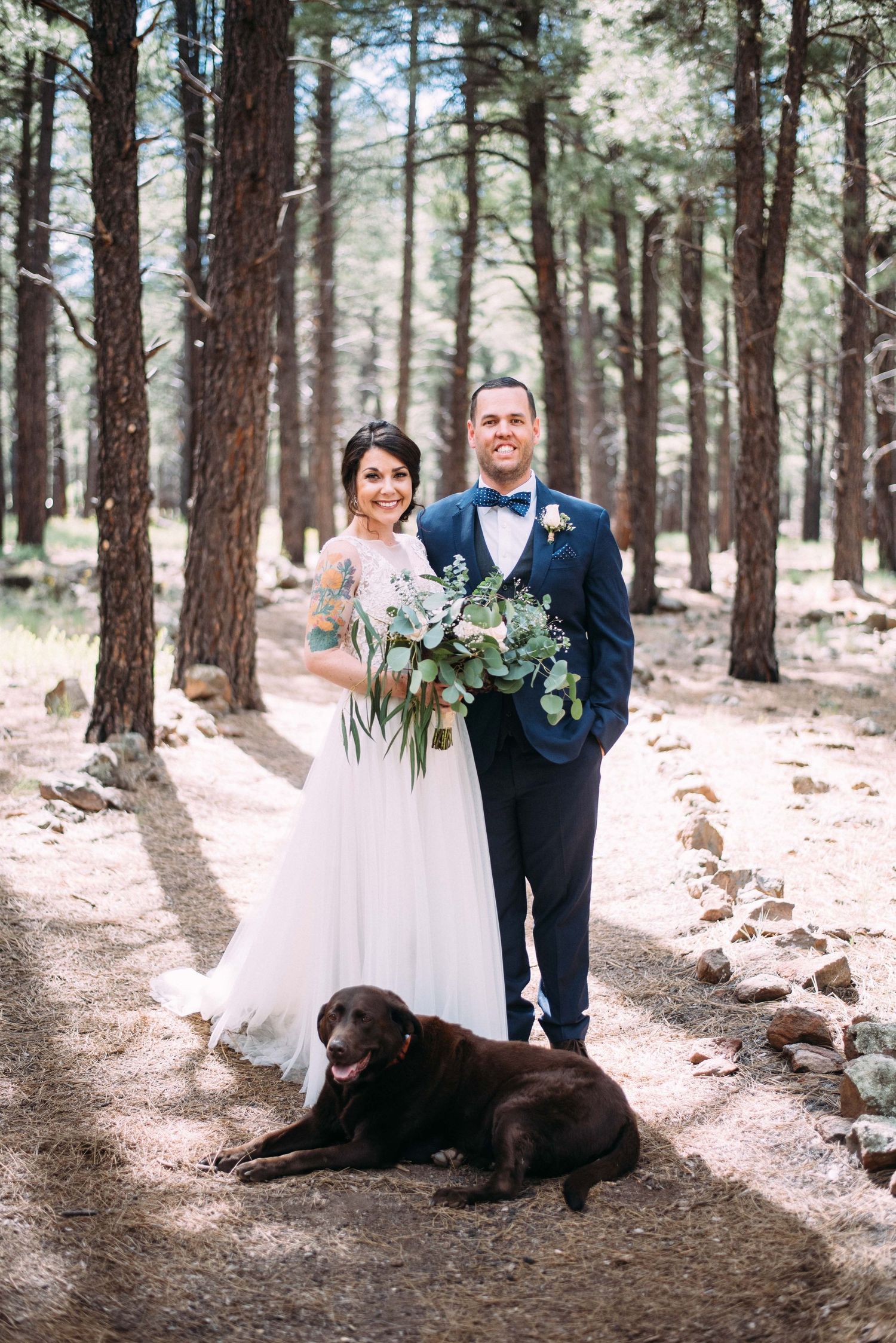 Wedding couple poses on forest path with black lab lying at their feet.
