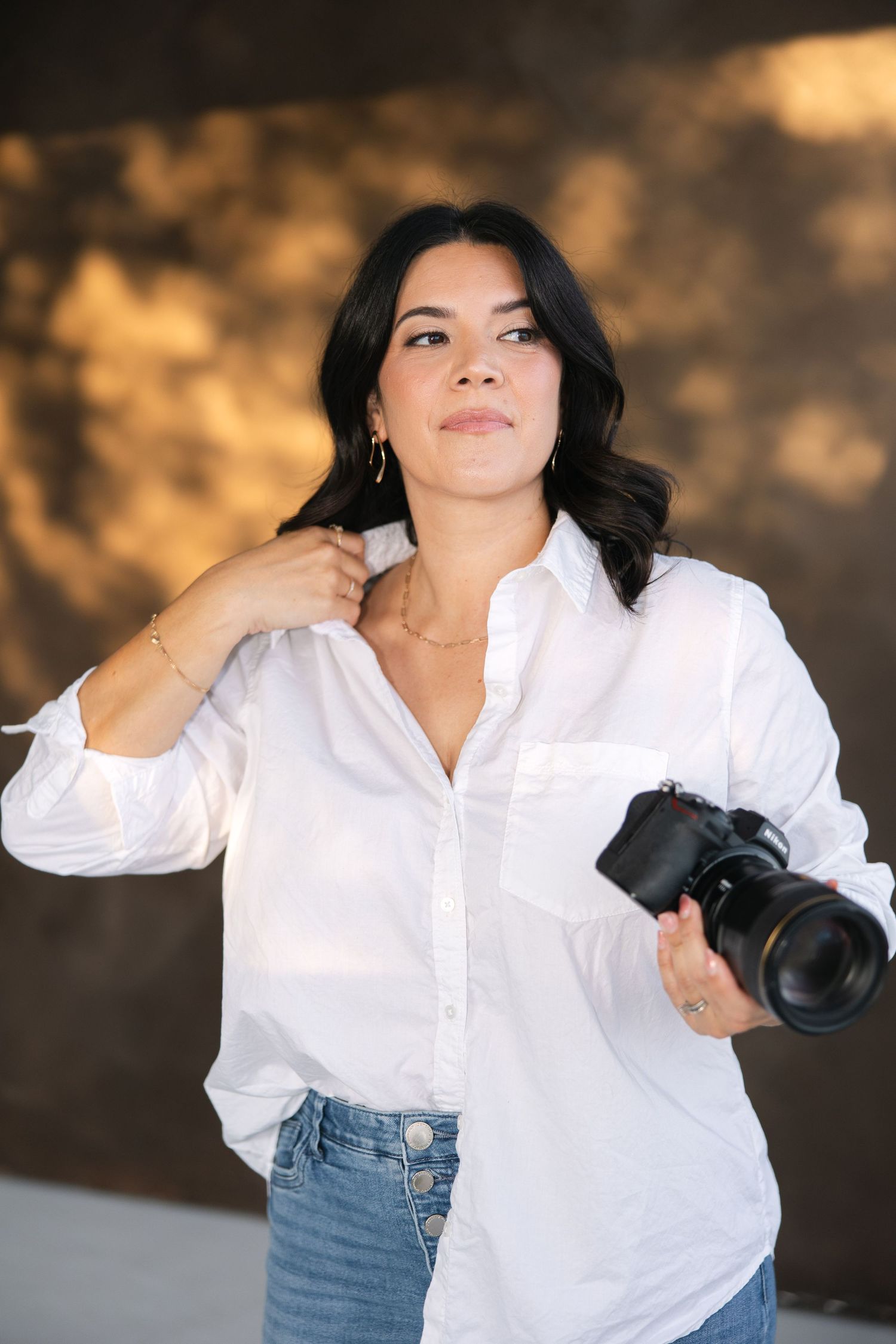 Photographer in white shirt and jeans posing with camera against dark background.