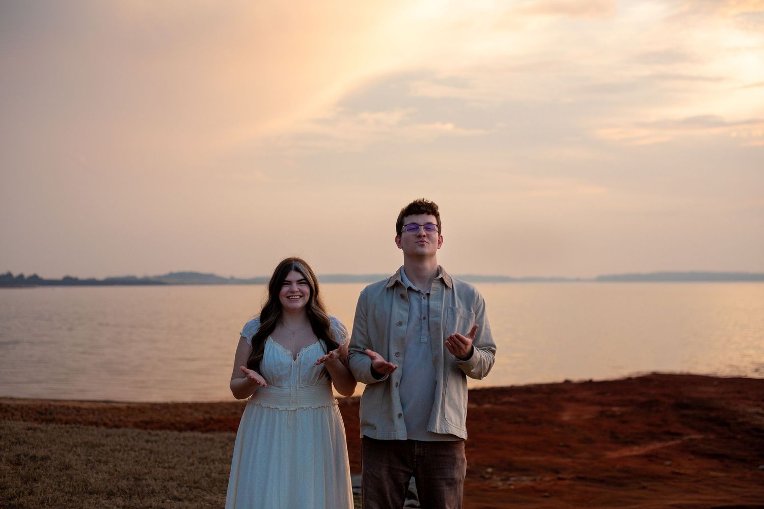A couple stands on a scenic overlook during golden hour at sunset with mountains and water in the background.