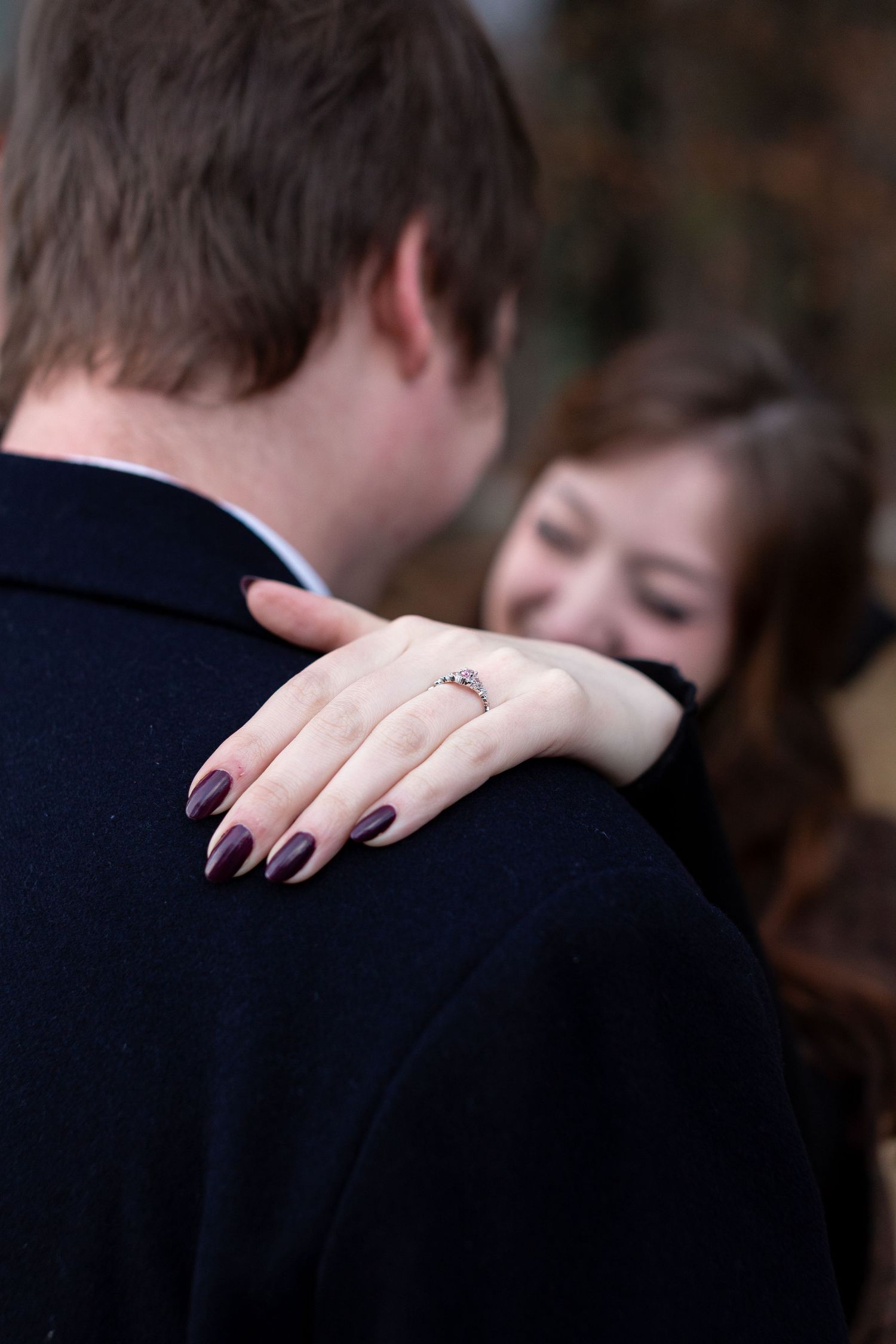 A close-up view of a woman's hand with dark nail polish resting on a man's shoulder in formal attire.
