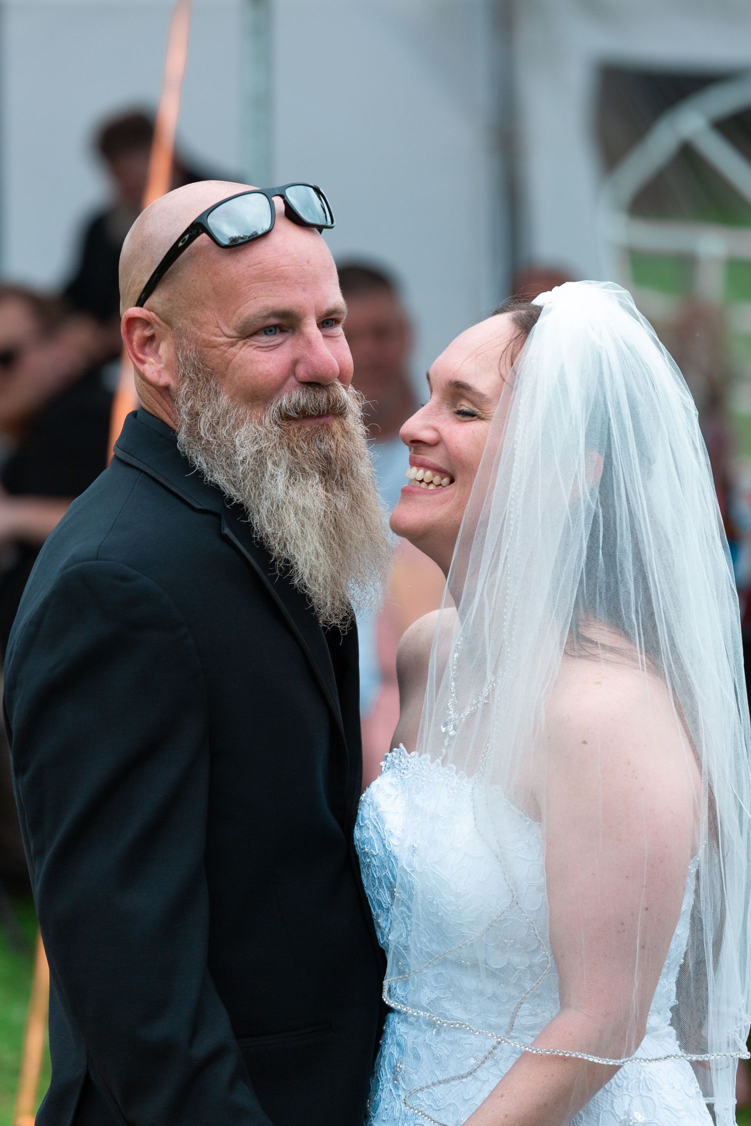 A joyful wedding couple sharing a candid moment outdoors with the groom sporting a long white beard and sunglasses.