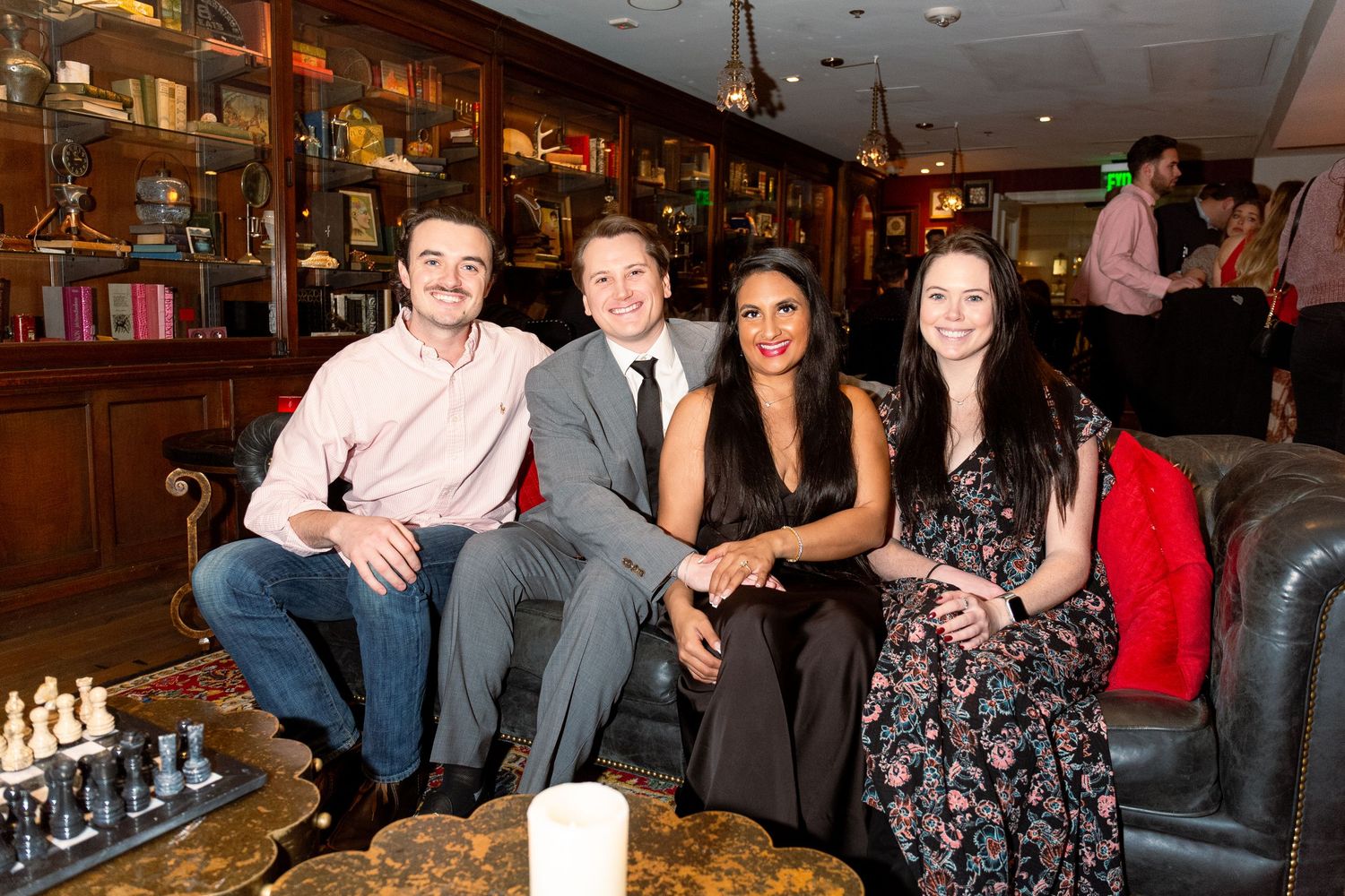 Four friends smiling together on a couch in an elegant bar with bookshelves and warm lighting.