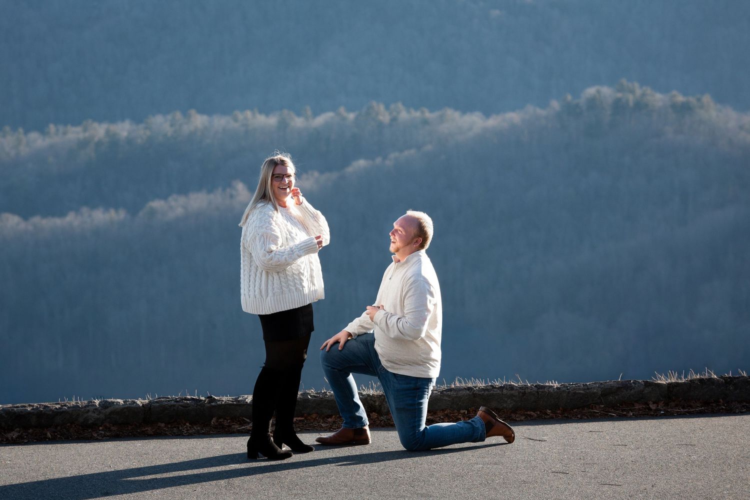 A romantic proposal scene unfolds on a concrete surface with mountains and cloudy skies in the background.
