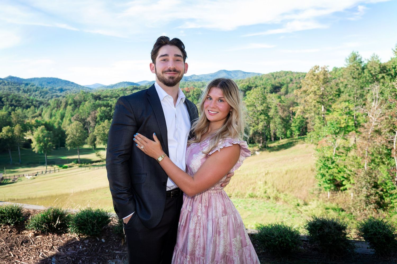 Smiling couple in formal wear embrace on a hillside overlook with green forest and mountains in the background on a sunny day.