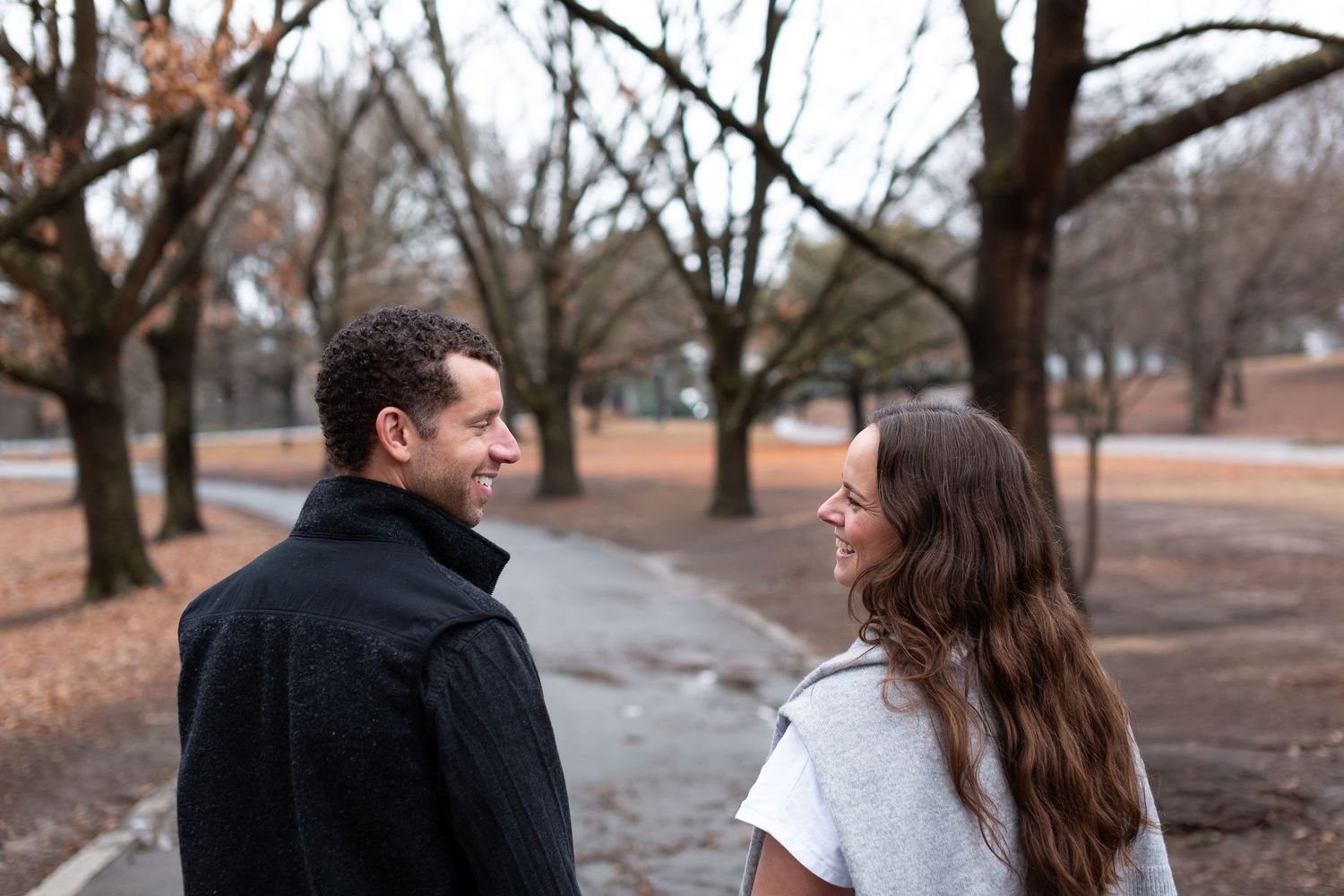 Two people stand facing each other on a path surrounded by bare trees in an autumn park setting.