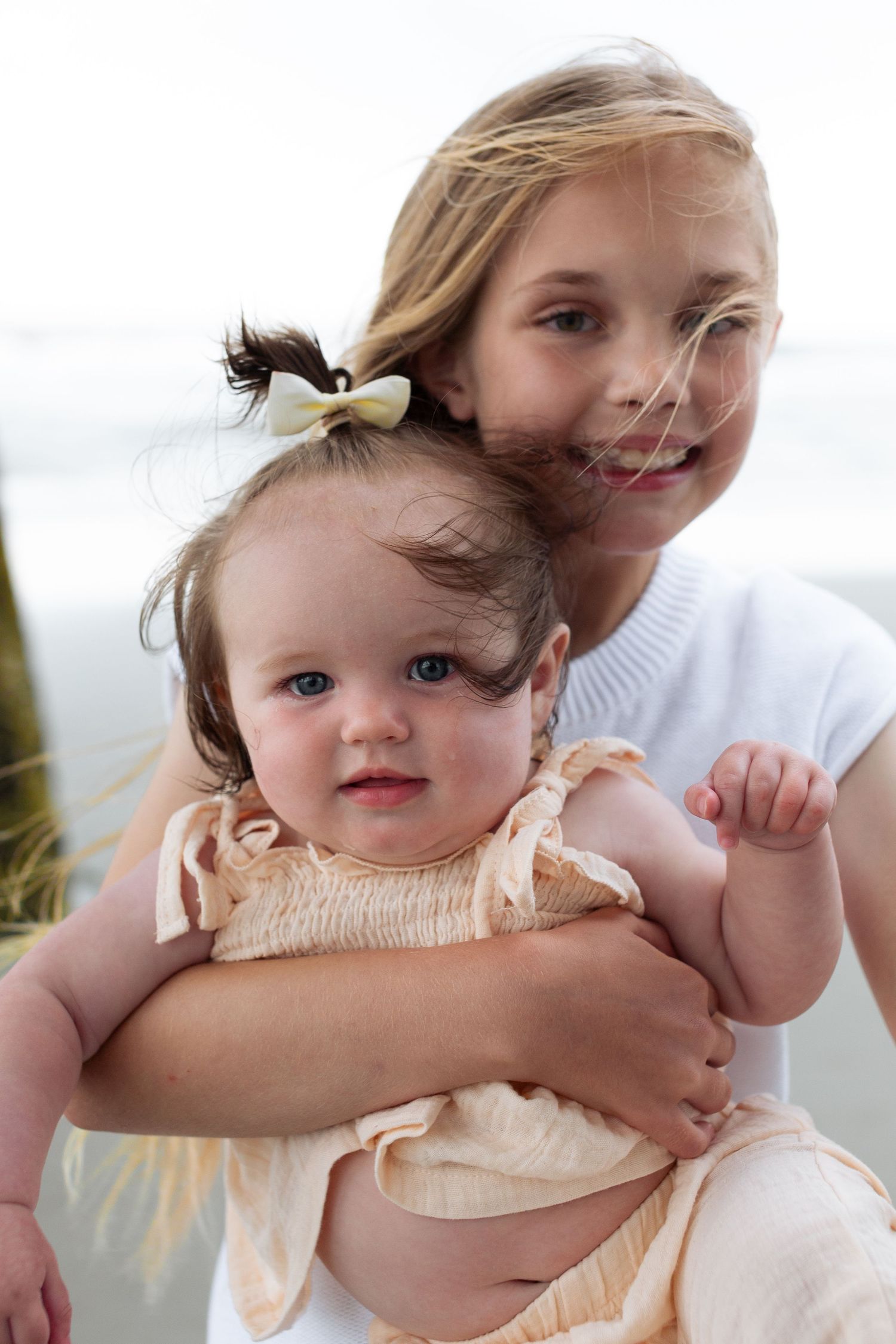 Two young children share a tender moment together outdoors, the older one holding the baby in a white dress.