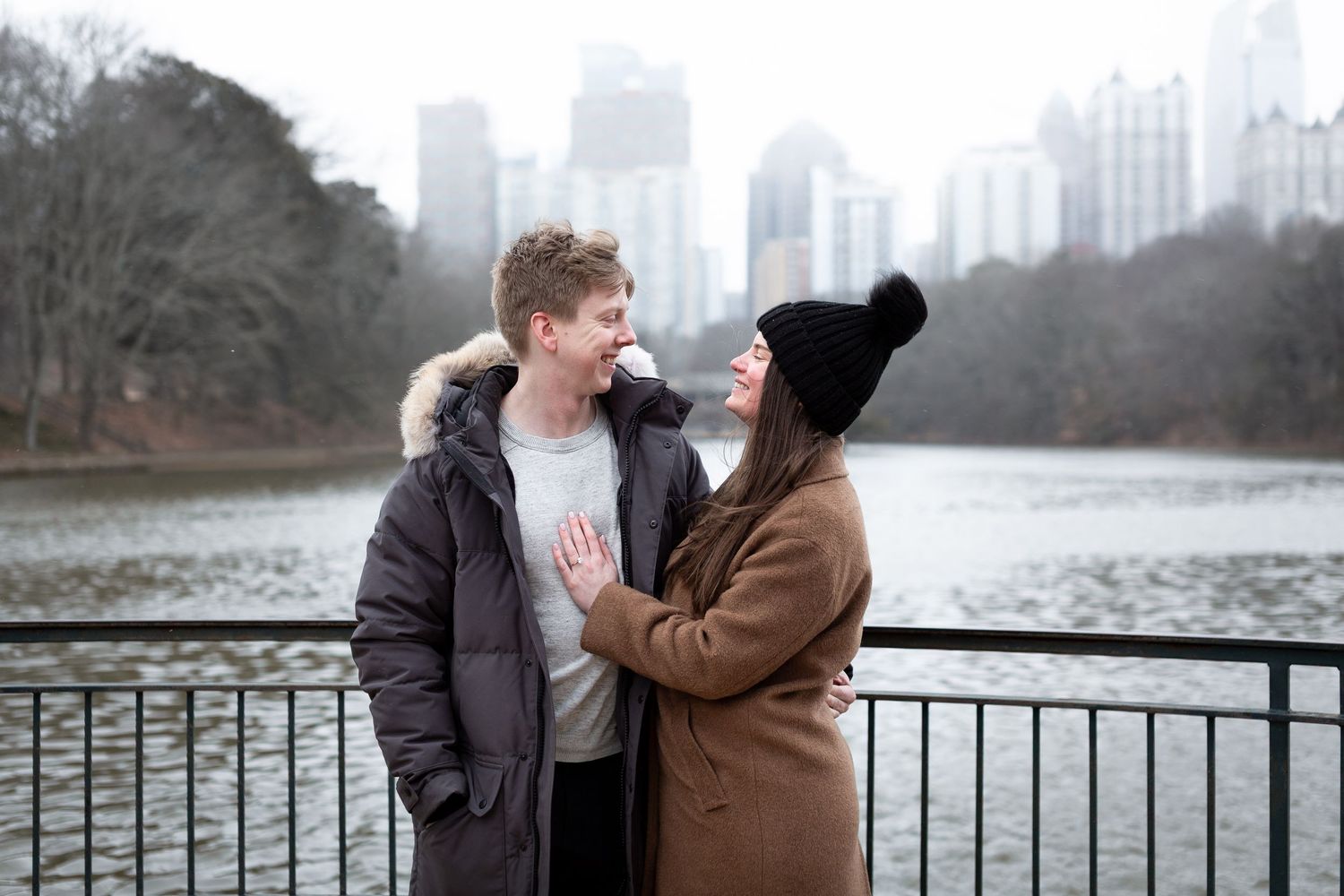 A couple shares tender moments along a lakeside railing with city skyline views in the background during winter.
