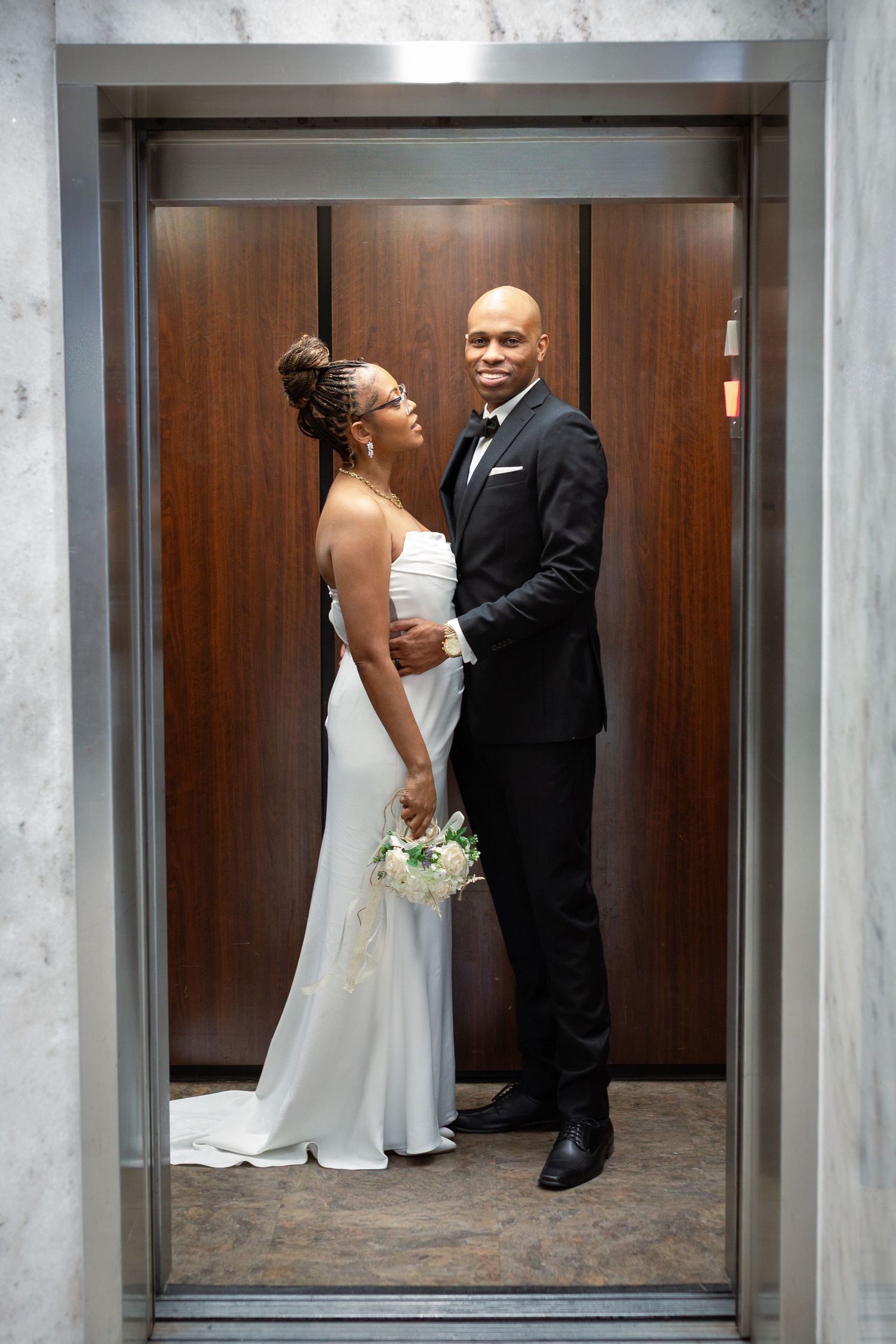 A couple in wedding attire shares a tender moment near an elevator in a modern building interior.