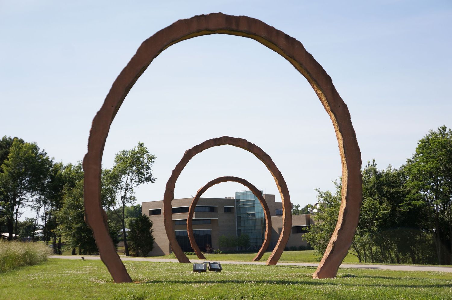 Large rust-colored metal arch sculptures on green lawn with modern building and trees in background on clear day.