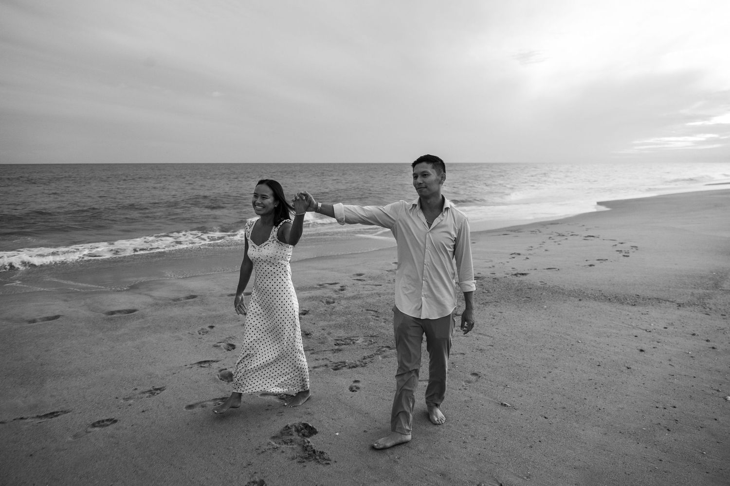 A black and white photo of a couple walking hand in hand along a beach at sunset.