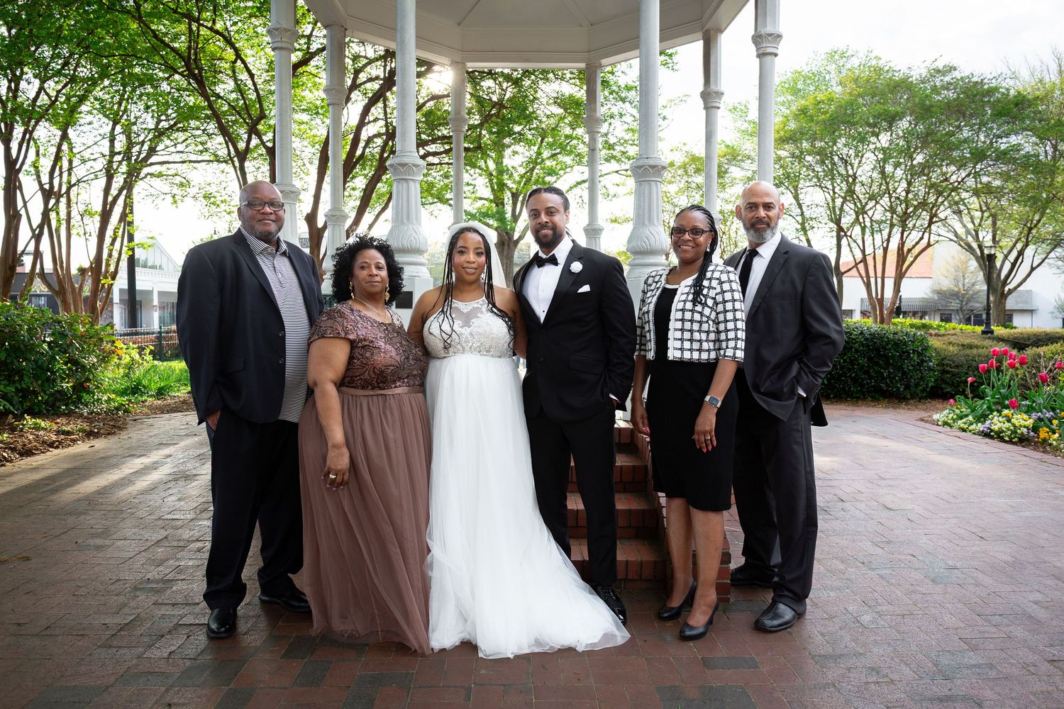 Wedding party of six posing under a gazebo in a lush garden, bride in white gown with groom in tuxedo surrounded by family.