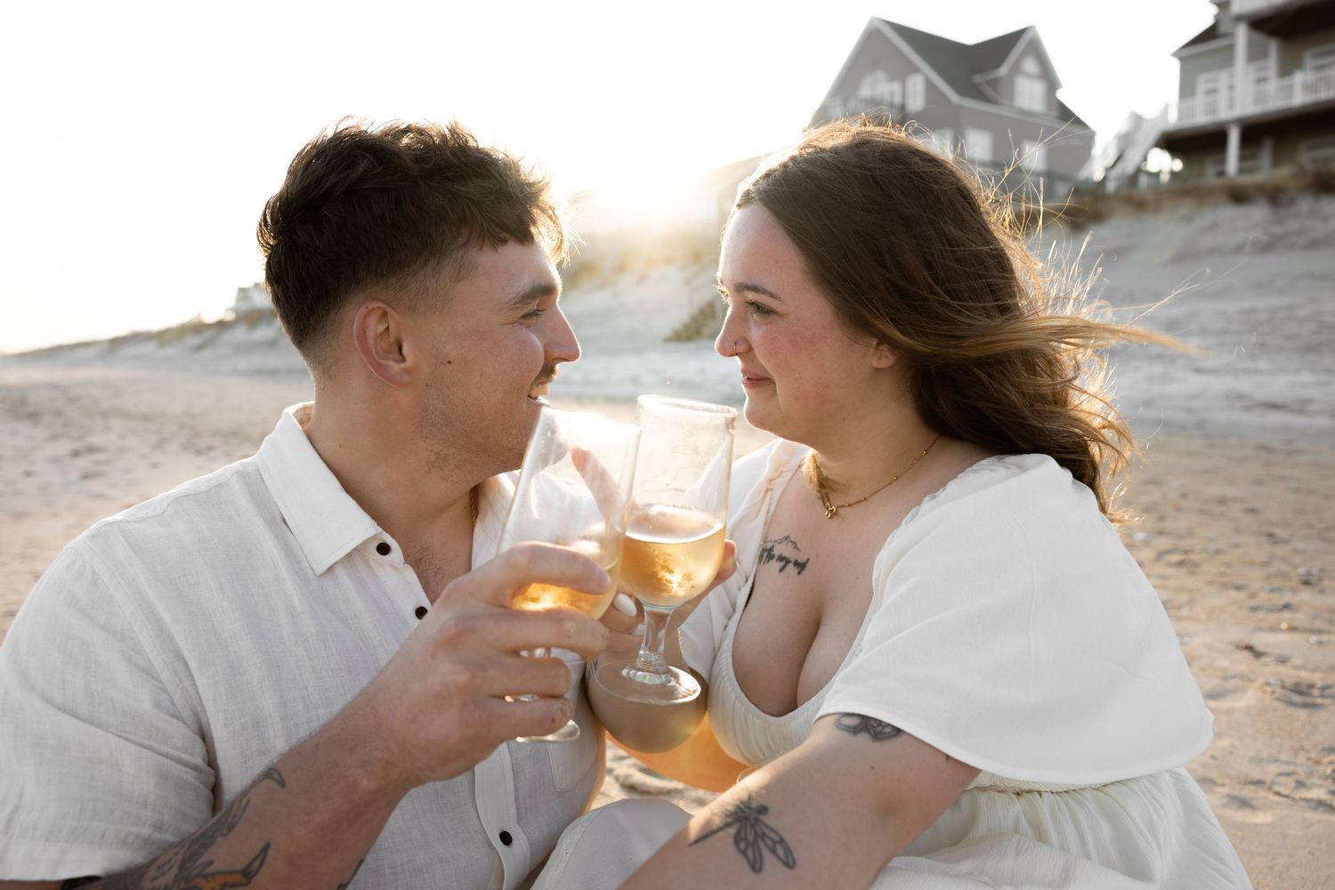 A couple shares an intimate moment with wine glasses on a sandy beach at sunset.