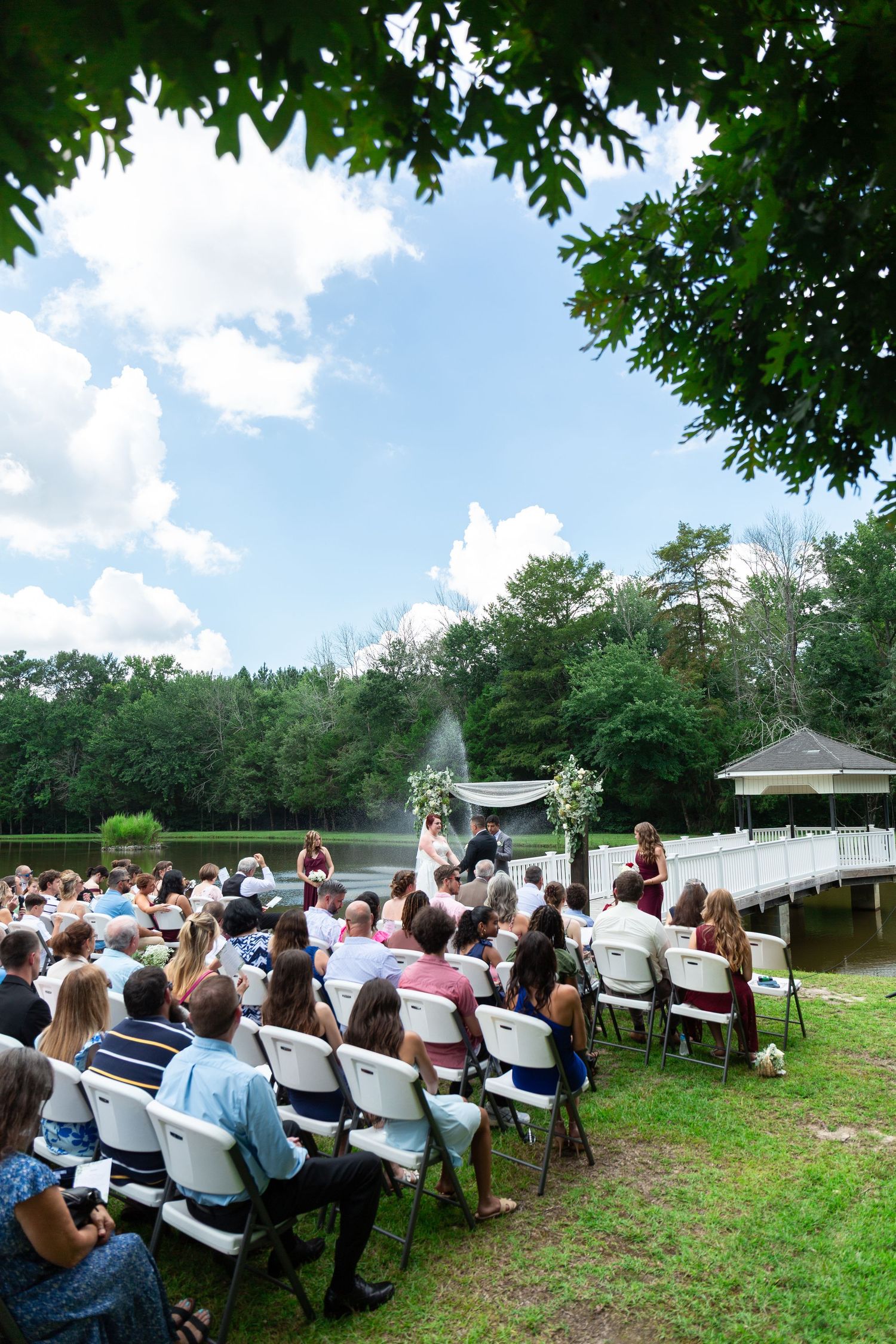 Outdoor wedding ceremony setup with white chairs arranged on grass under trees and blue sky.