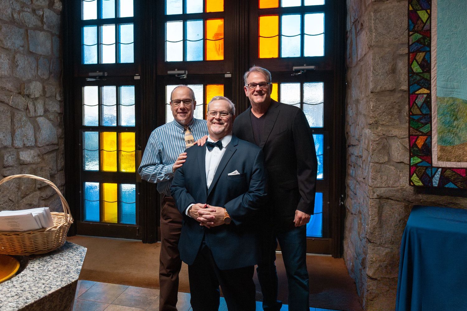 Wedding party gathers in front of stained glass windows inside a stone church building.
