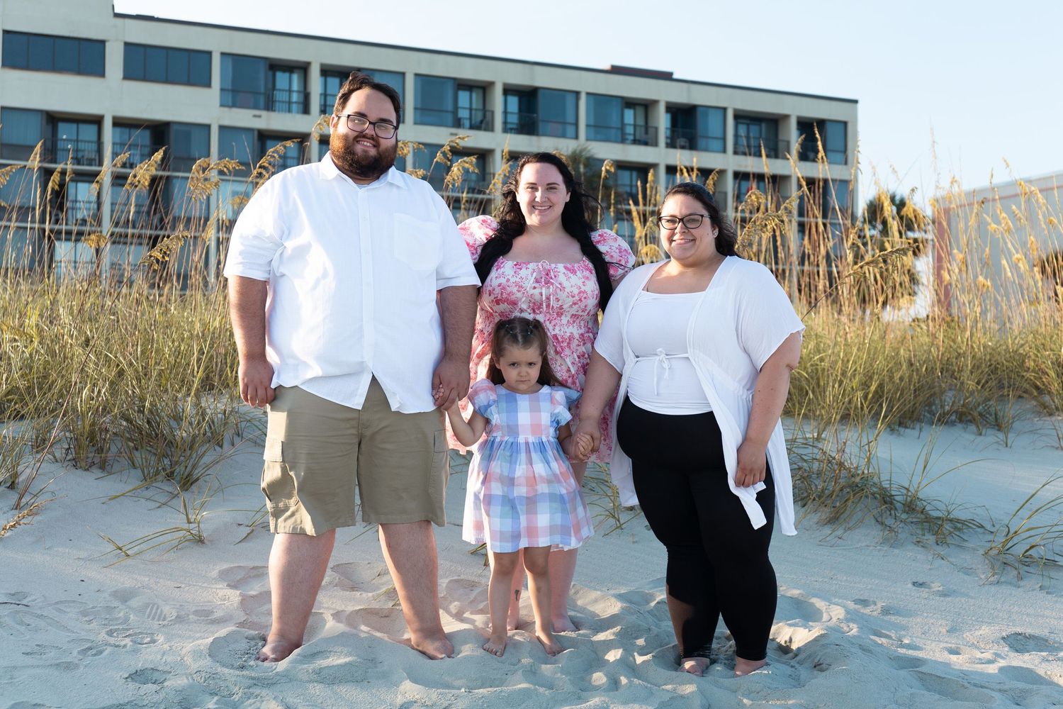 Family group poses together on sandy beach with dunes and buildings in background.
