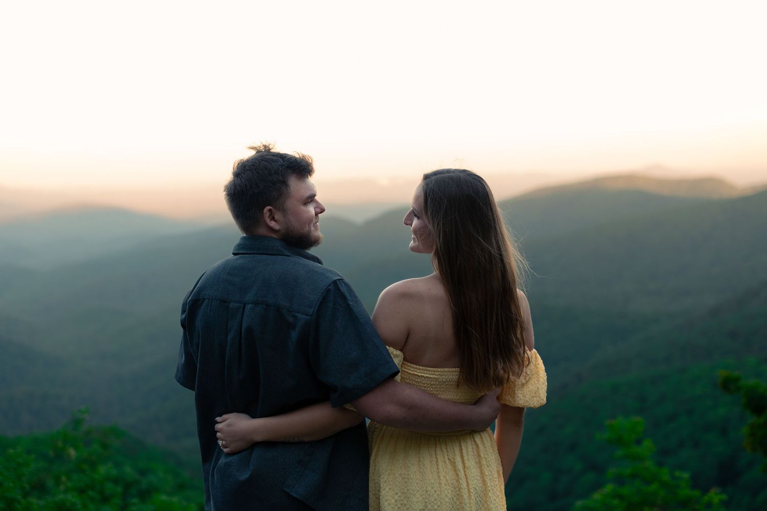 Couple embracing at sunset overlooking misty green mountain landscape, woman in yellow dress, man in dark shirt.