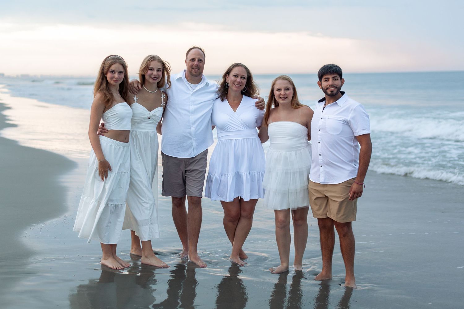 A group wearing coordinated white outfits poses together on a beach at sunset.