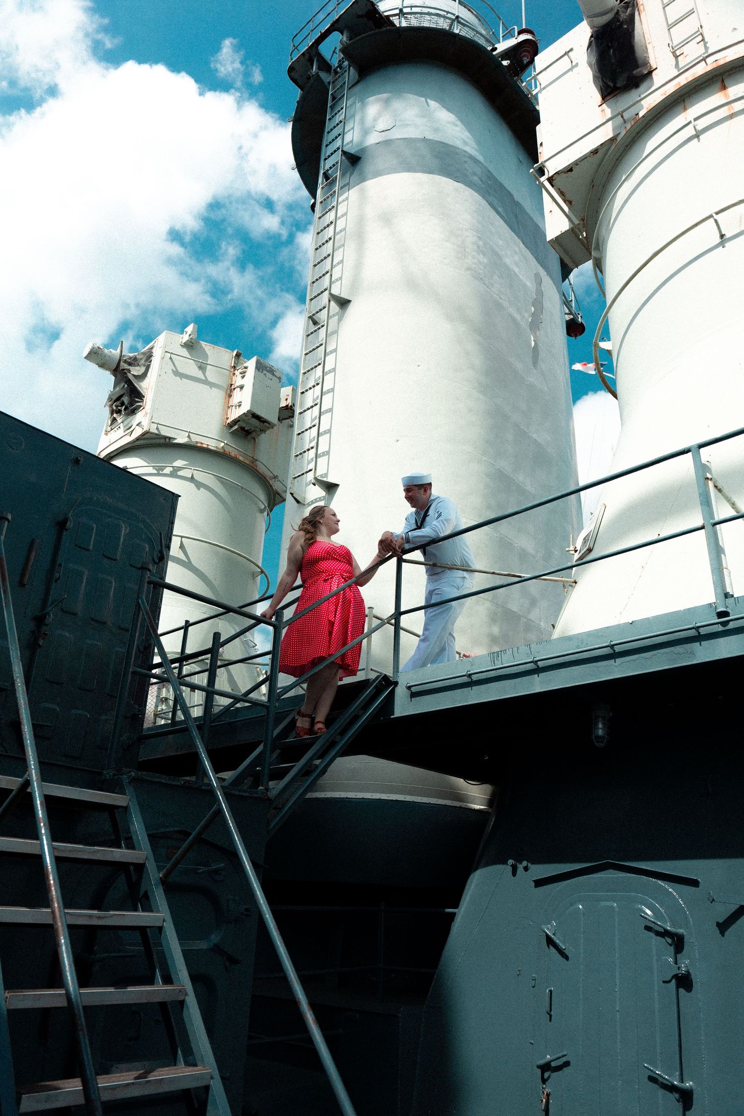 People climb white metal ladders on a ship's exterior under a bright blue sky with white clouds.