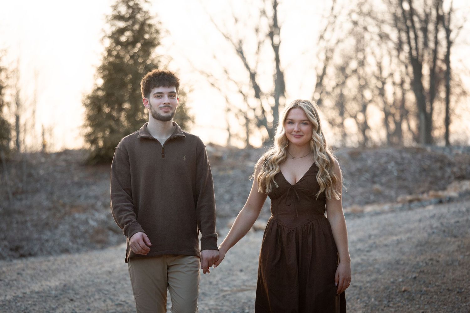 A couple holds hands while walking on a dirt path at sunset with bare trees in the background.