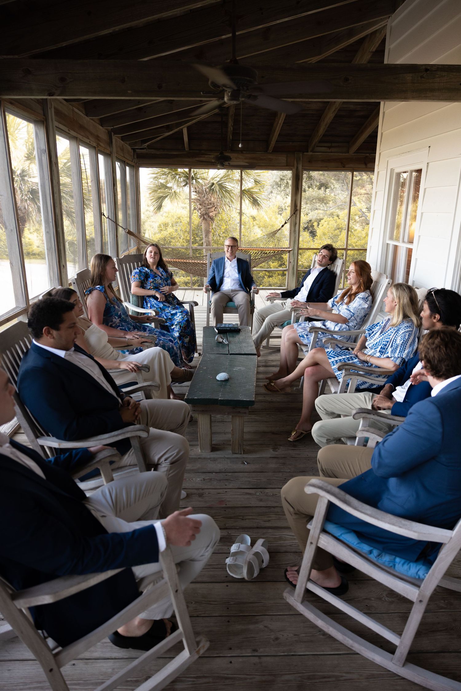 Group of people relaxing in white rocking chairs on a rustic wooden porch during a sunny afternoon.