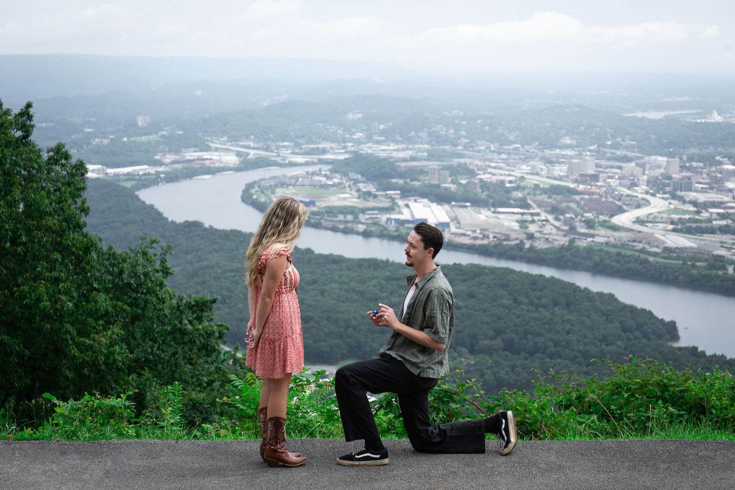 Man kneeling on overlook proposing to woman with scenic river valley and city views in background.