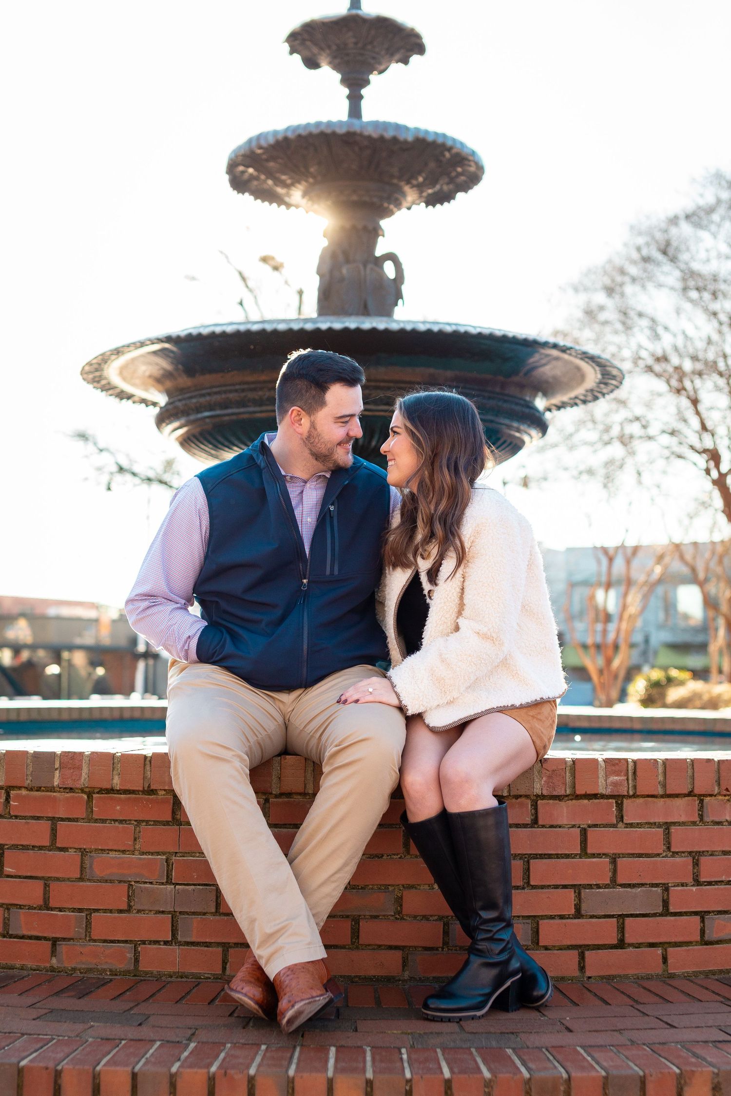 A couple sits together on a brick ledge near an ornate fountain during a romantic outdoor photoshoot.