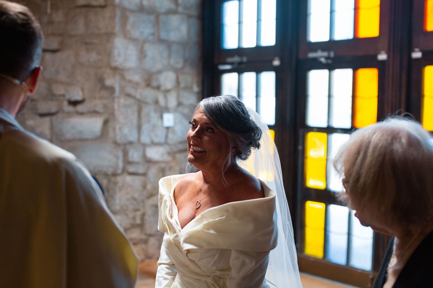 A group of people in formal attire gather around a table in a rustic stone-walled room with yellow-lit windows.