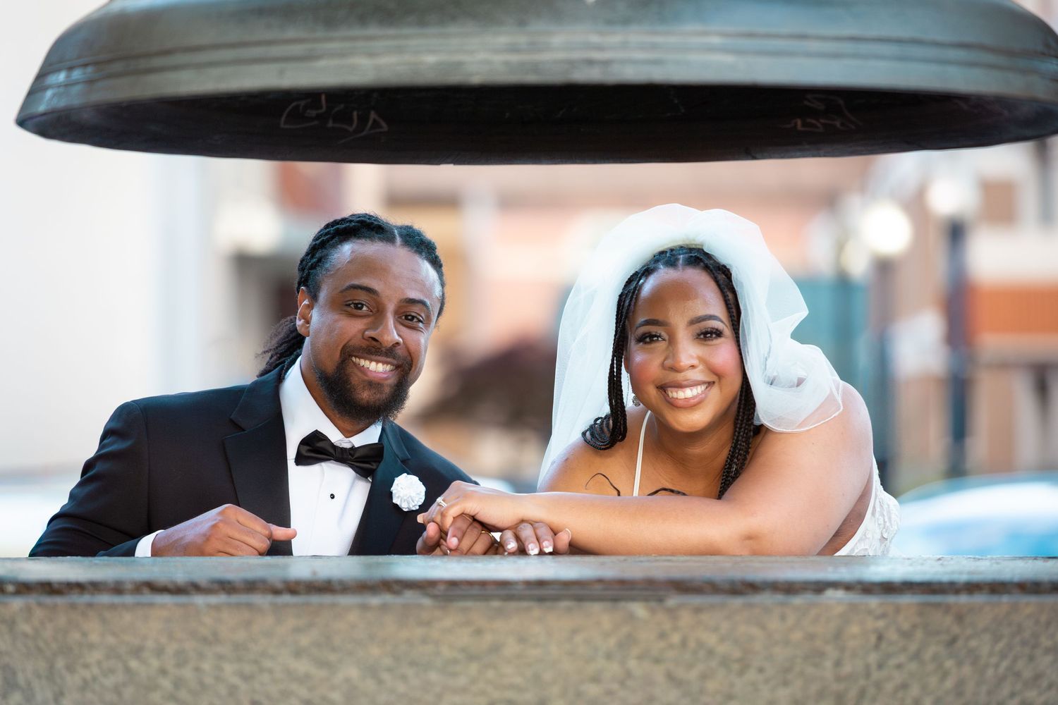 A smiling bride and groom in wedding attire pose together near a large decorative bell outdoors.