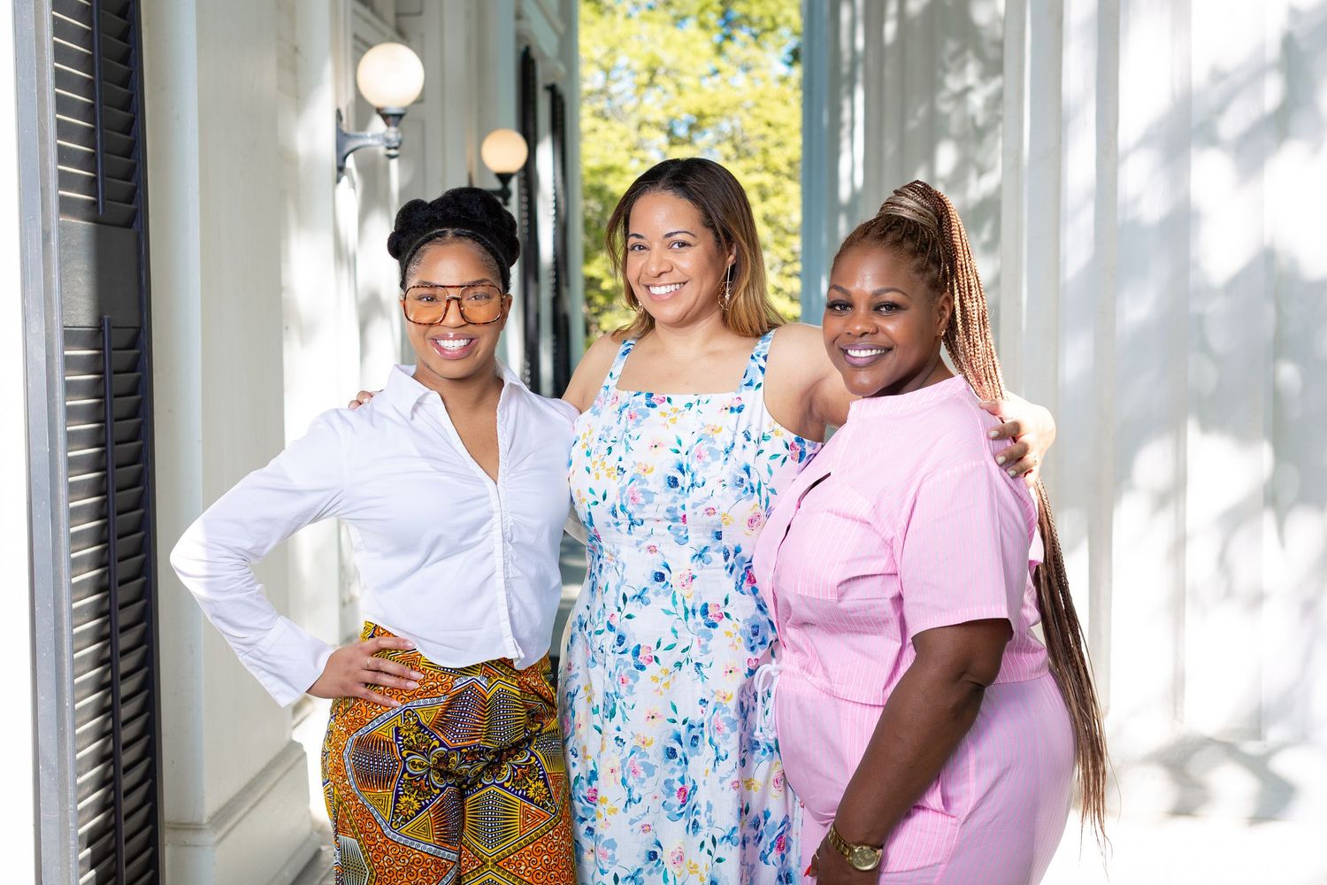 Three women smiling and posing together outdoors, wearing colorful outfits in a bright, sunlit setting.