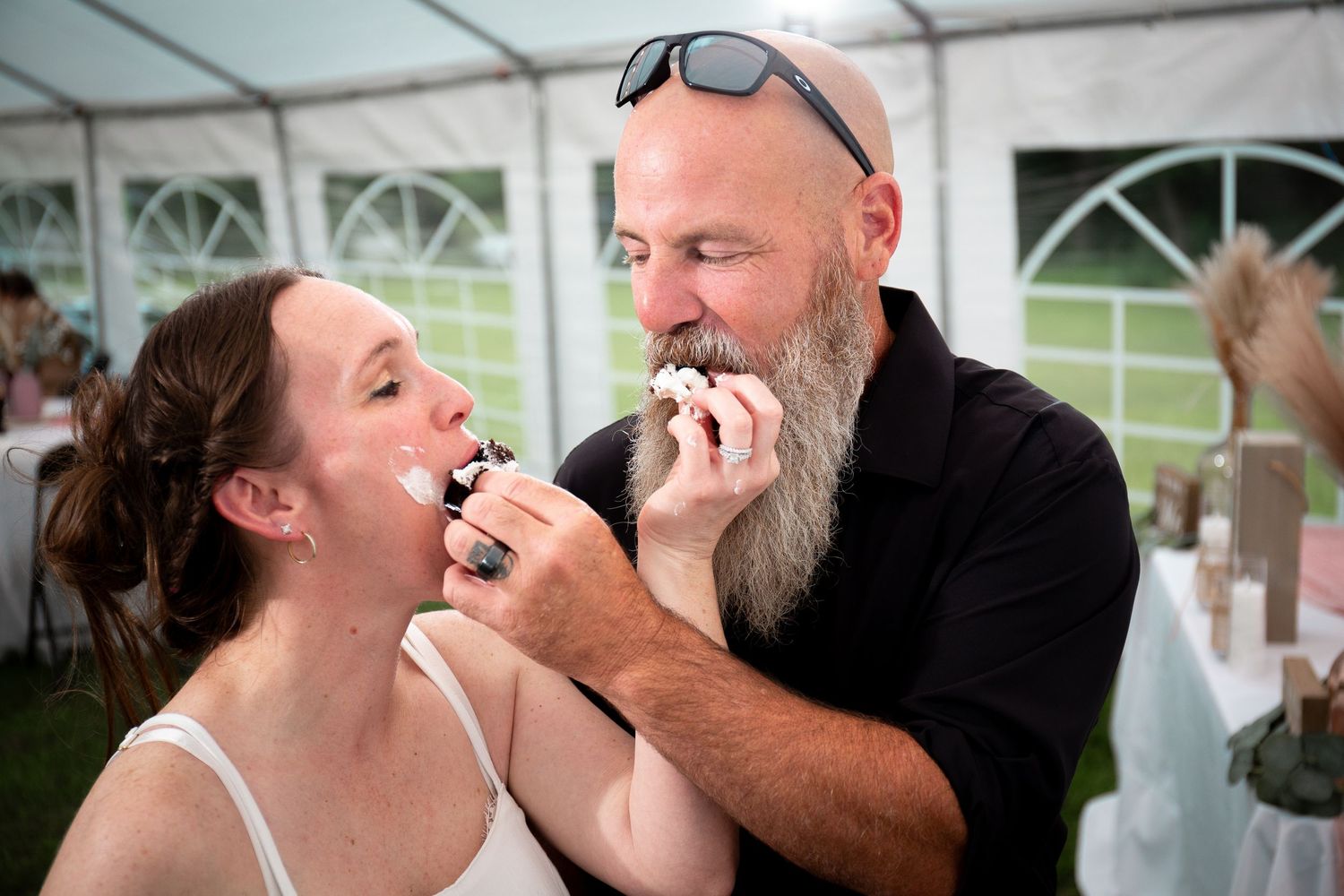A couple shares a playful moment while eating cake together at a wedding reception under a white tent.
