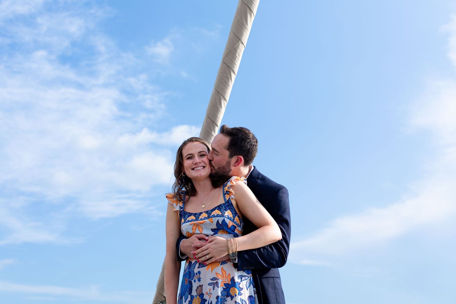 A romantic couple embraces against a bright blue sky with a pole in the background.