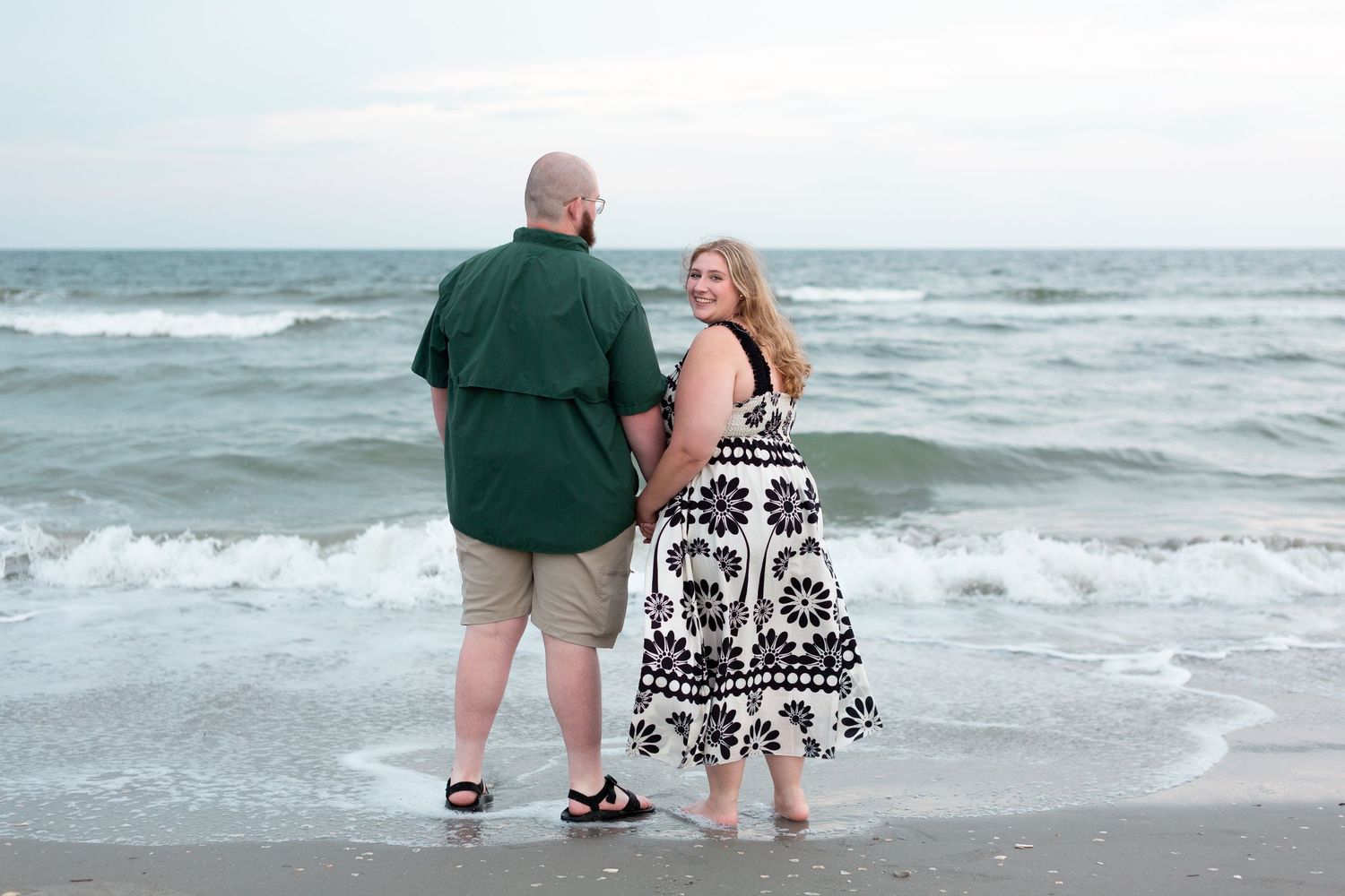 Couple walking along beach shoreline holding hands as waves crash on sandy coast at sunset.