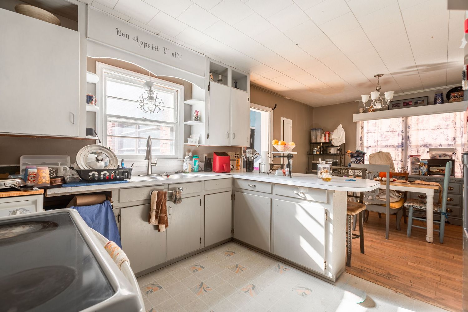 A bright L-shaped kitchen with white cabinets, wooden floors, and multiple windows providing natural illumination.