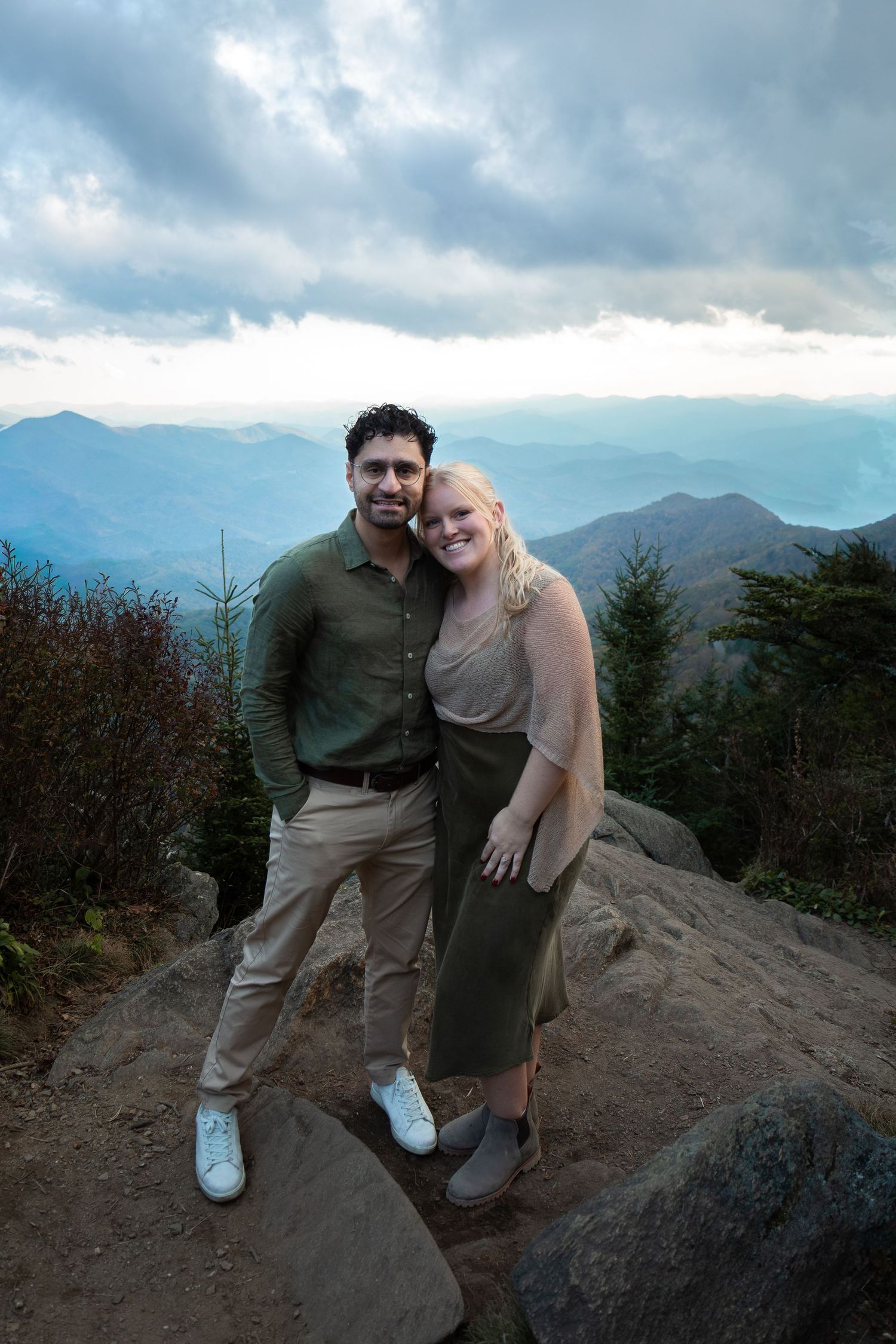 A smiling couple poses together on a rocky mountain overlook with layered blue ridges and a cloudy sky behind them.