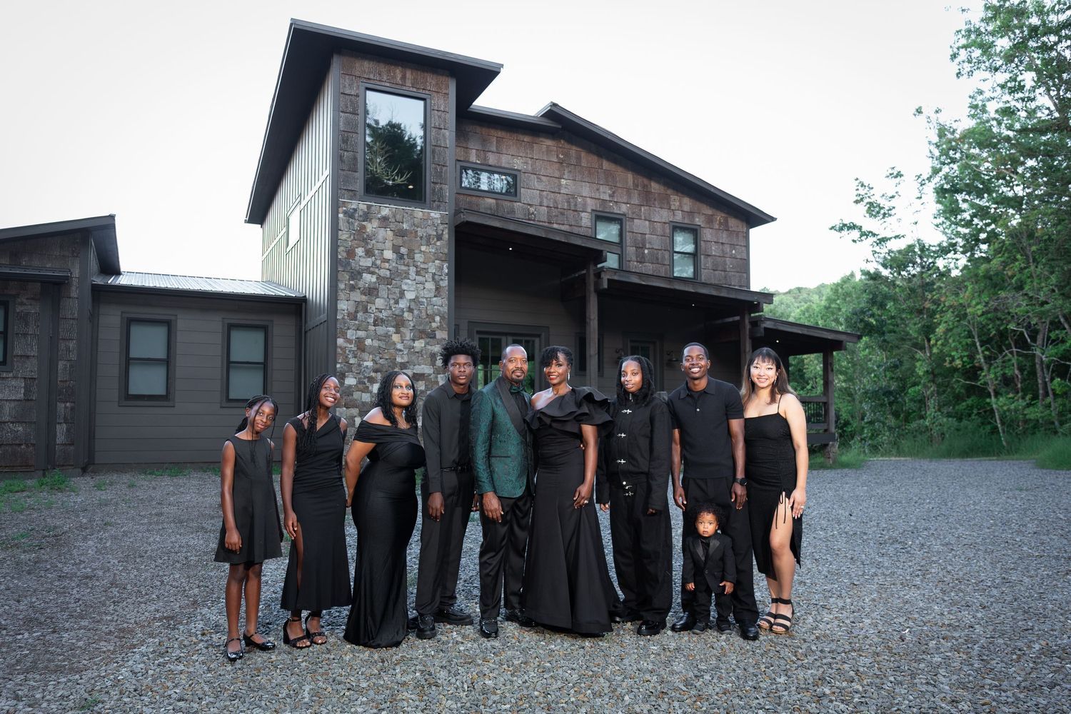 A group of twelve people dressed in black stand together on a gravel driveway in front of a modern stone and wood house surrounded by trees.