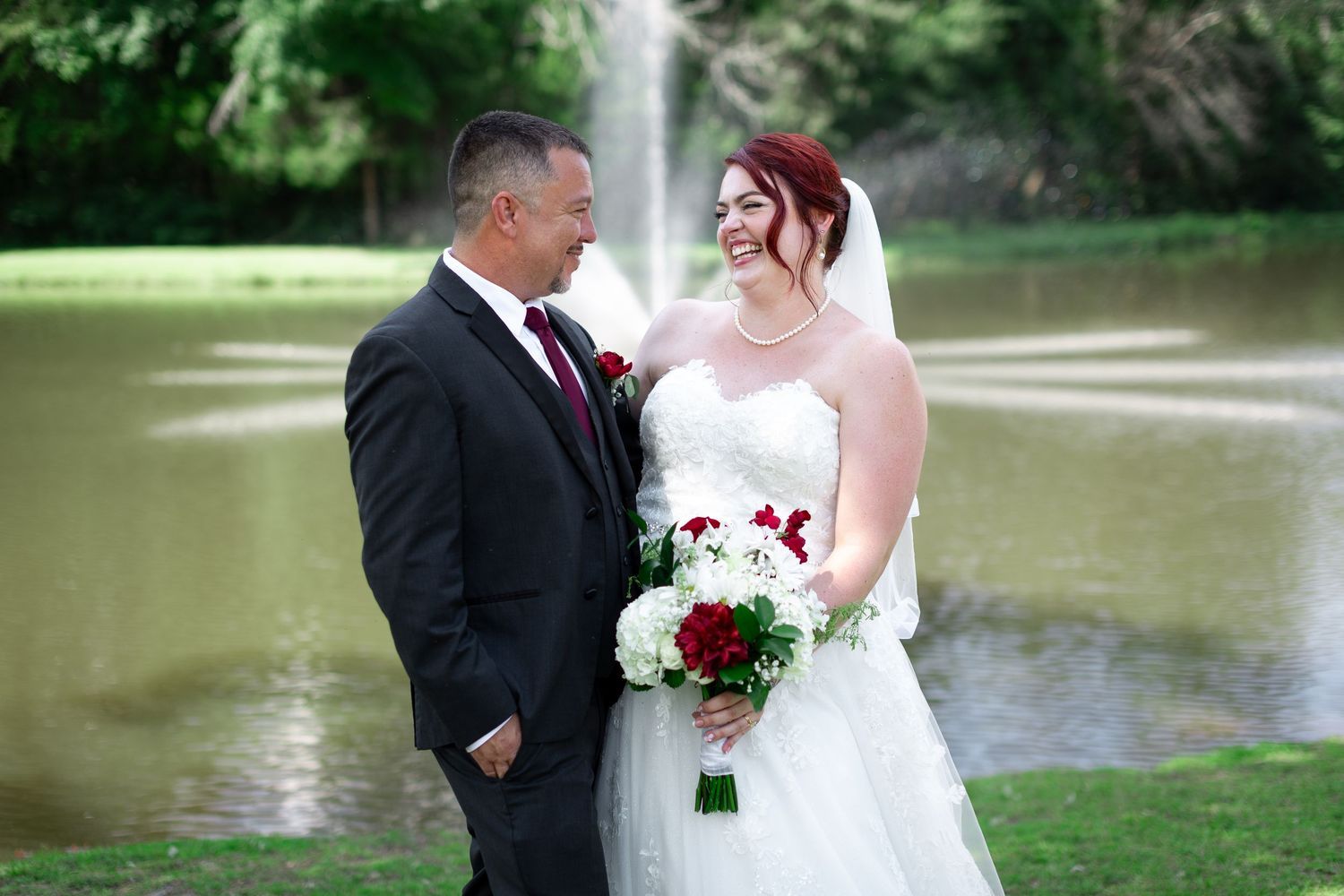A smiling bride in a white gown and veil holds a red and white bouquet while laughing with her groom in a dark suit beside a fountain pond.