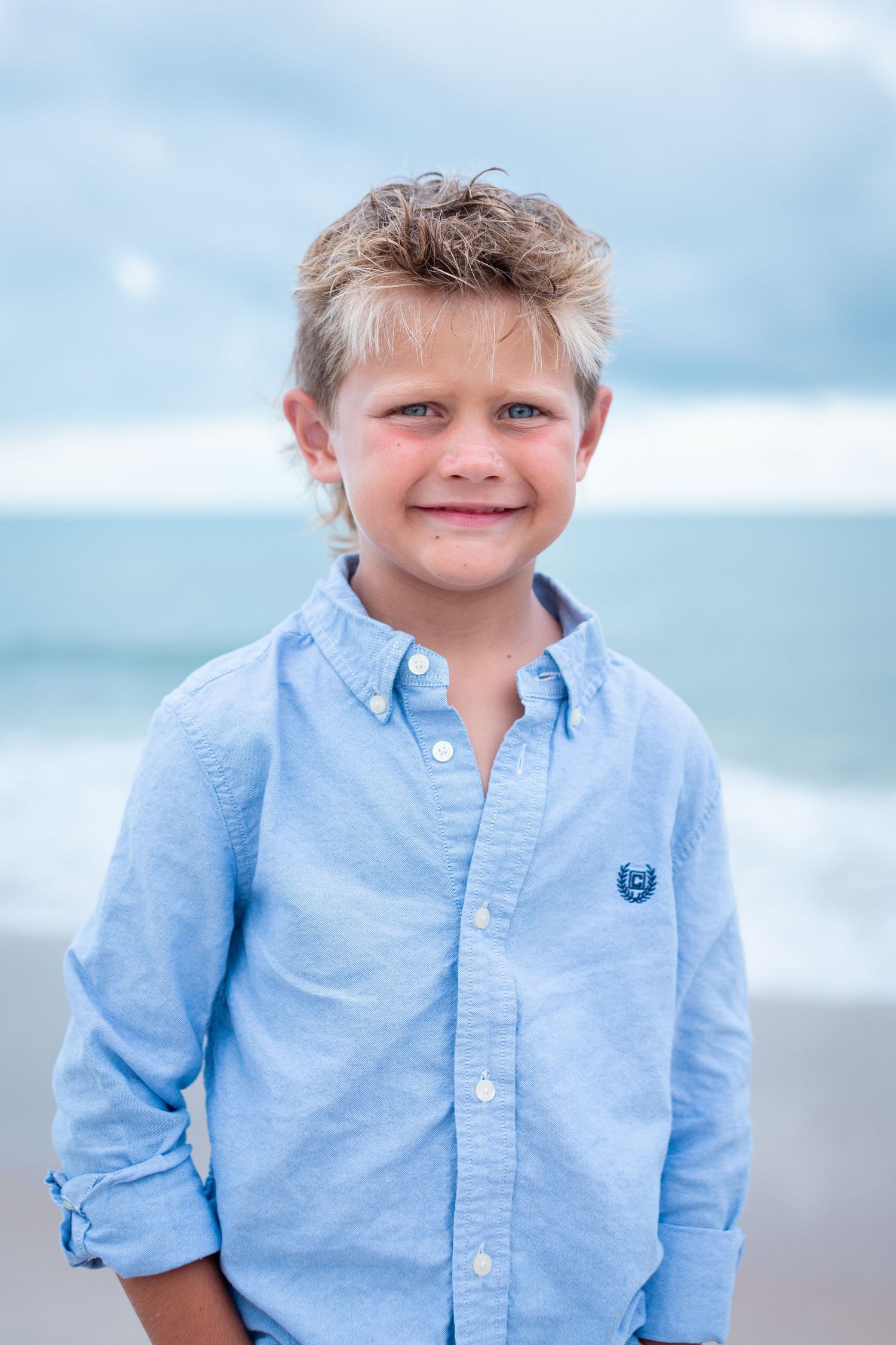 Young child in light blue button-down shirt poses on beach with ocean waves in background.