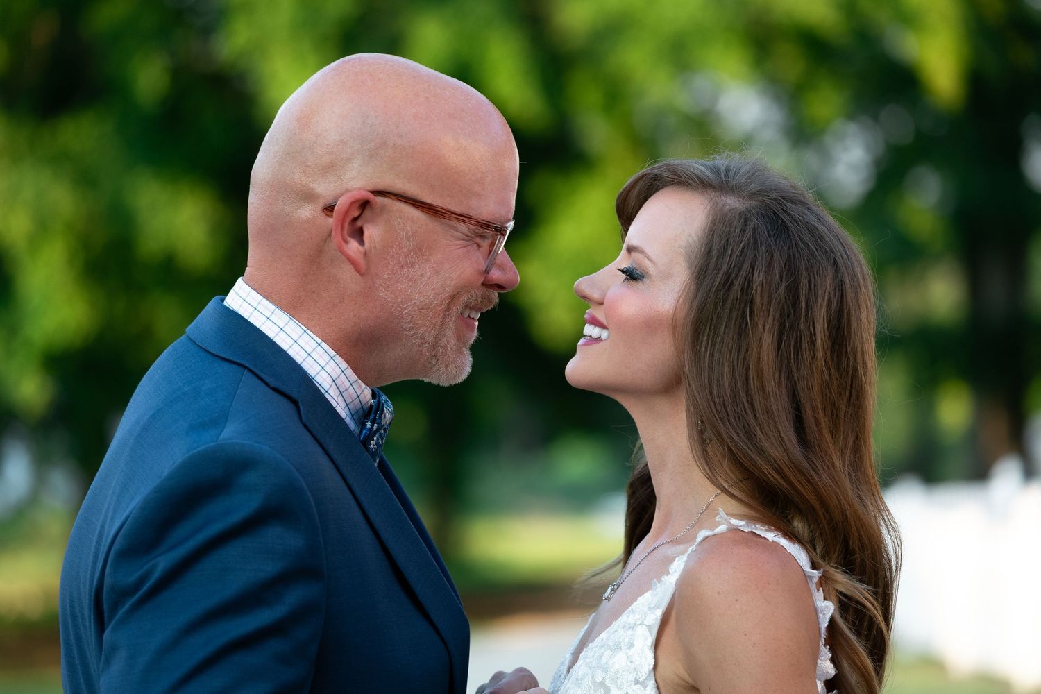 Smiling bride in a white dress gazes lovingly at her bald, bespectacled groom in a navy suit during an outdoor wedding portrait.