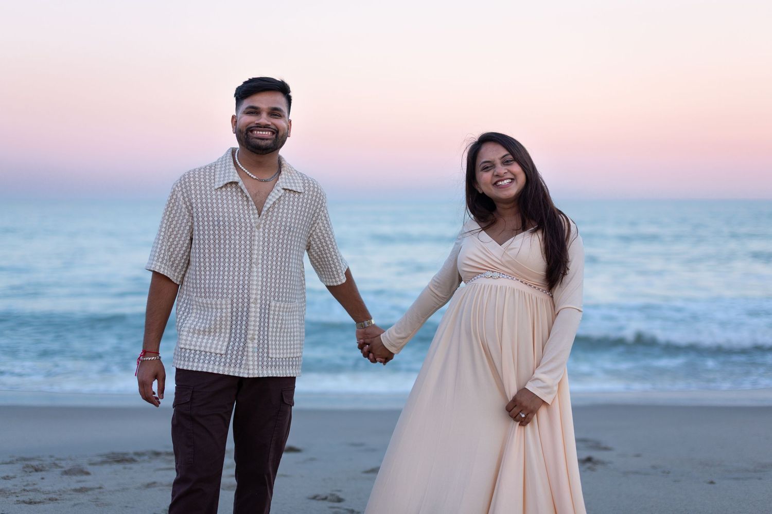 A smiling couple holds hands on the beach at sunset, the woman in a flowing dress showing her baby bump.