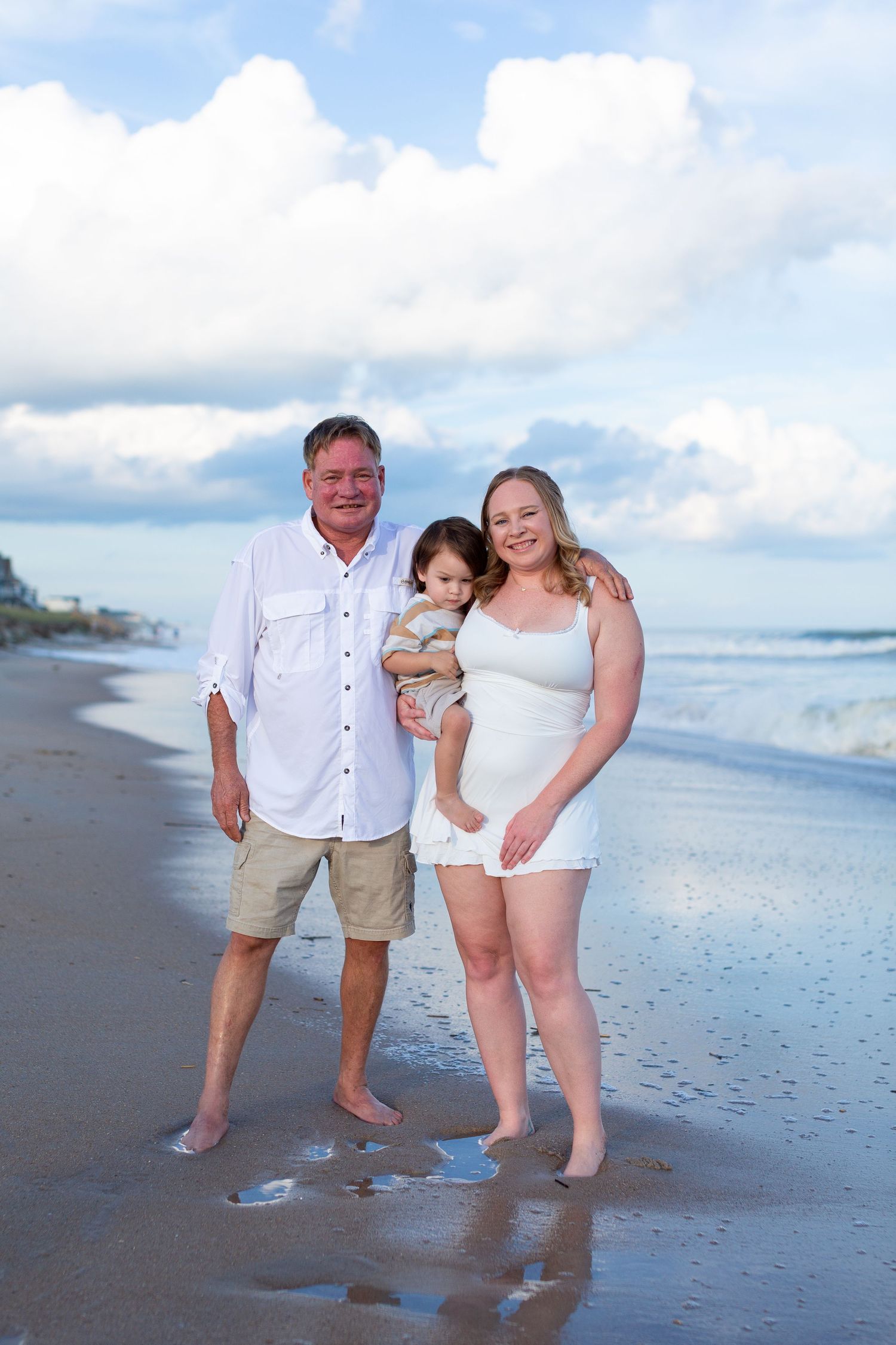 Family of three poses on sandy beach shoreline with ocean waves, wearing white outfits, holding toddler under cloudy blue sky.