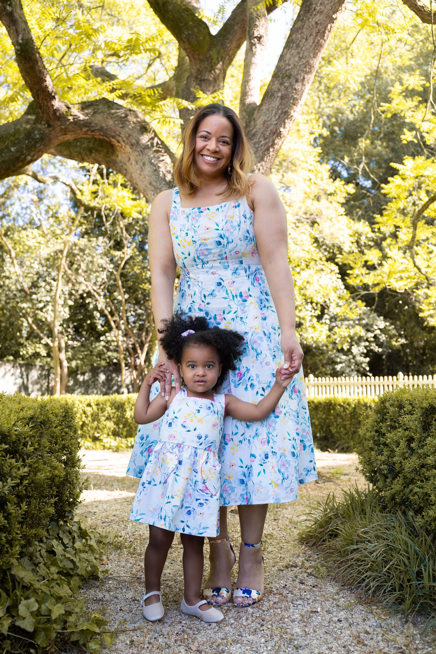 Mother and daughter in matching floral dresses pose together in a sunny garden with lush green trees.
