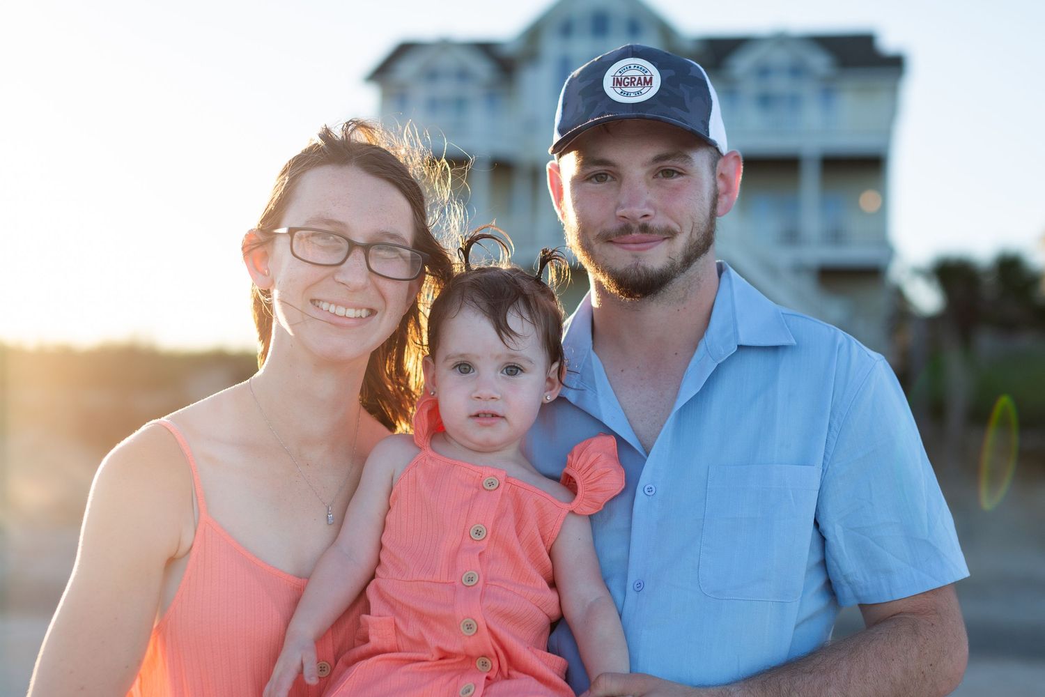 A family of three poses together at sunset with beach houses visible in the background.