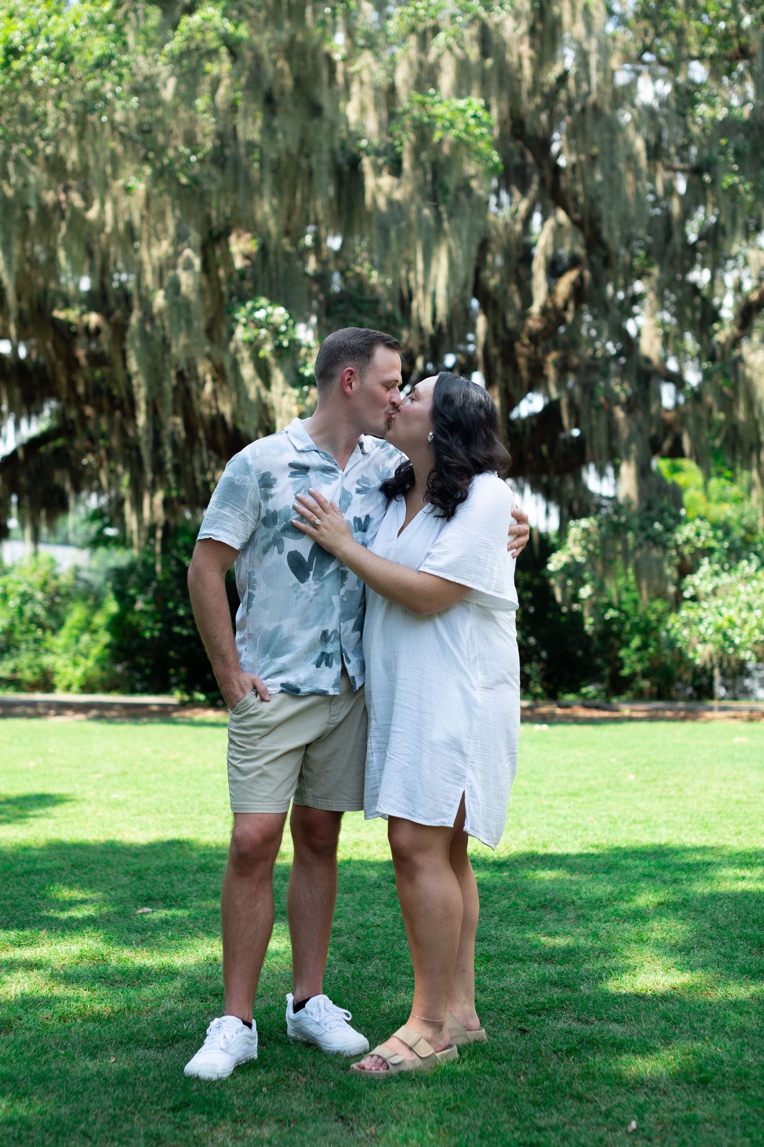 A couple in matching white outfits poses playfully on a lush green lawn with Spanish moss-draped trees in the background.