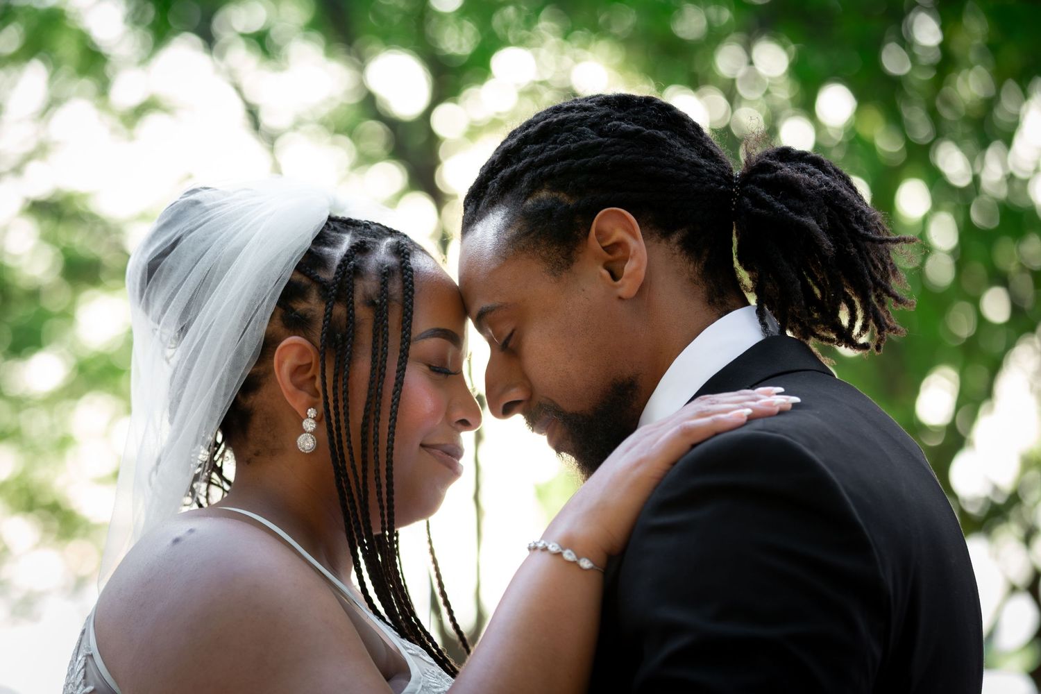 A bride and groom in formal wedding attire share an intimate forehead-to-forehead moment outdoors.